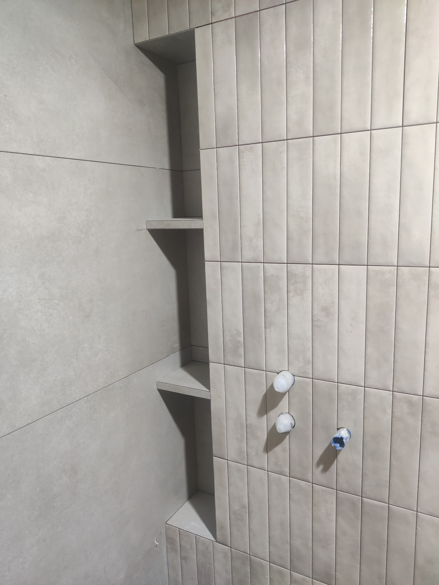 Wykończona łazienka z szarymi, pionowo ułożonymi płytkami i wnękami z półkami. Widoczne przygotowane punkty wodne. Minimalistyczny design.