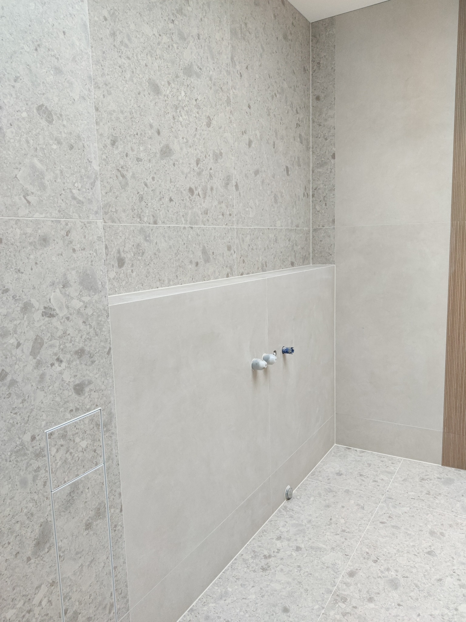 Jasnoszara łazienka z płytkami imitującymi kamień, widoczne przyłącza wodne i odpływ liniowy. Minimalistyczny design, neutralna kolorystyka.