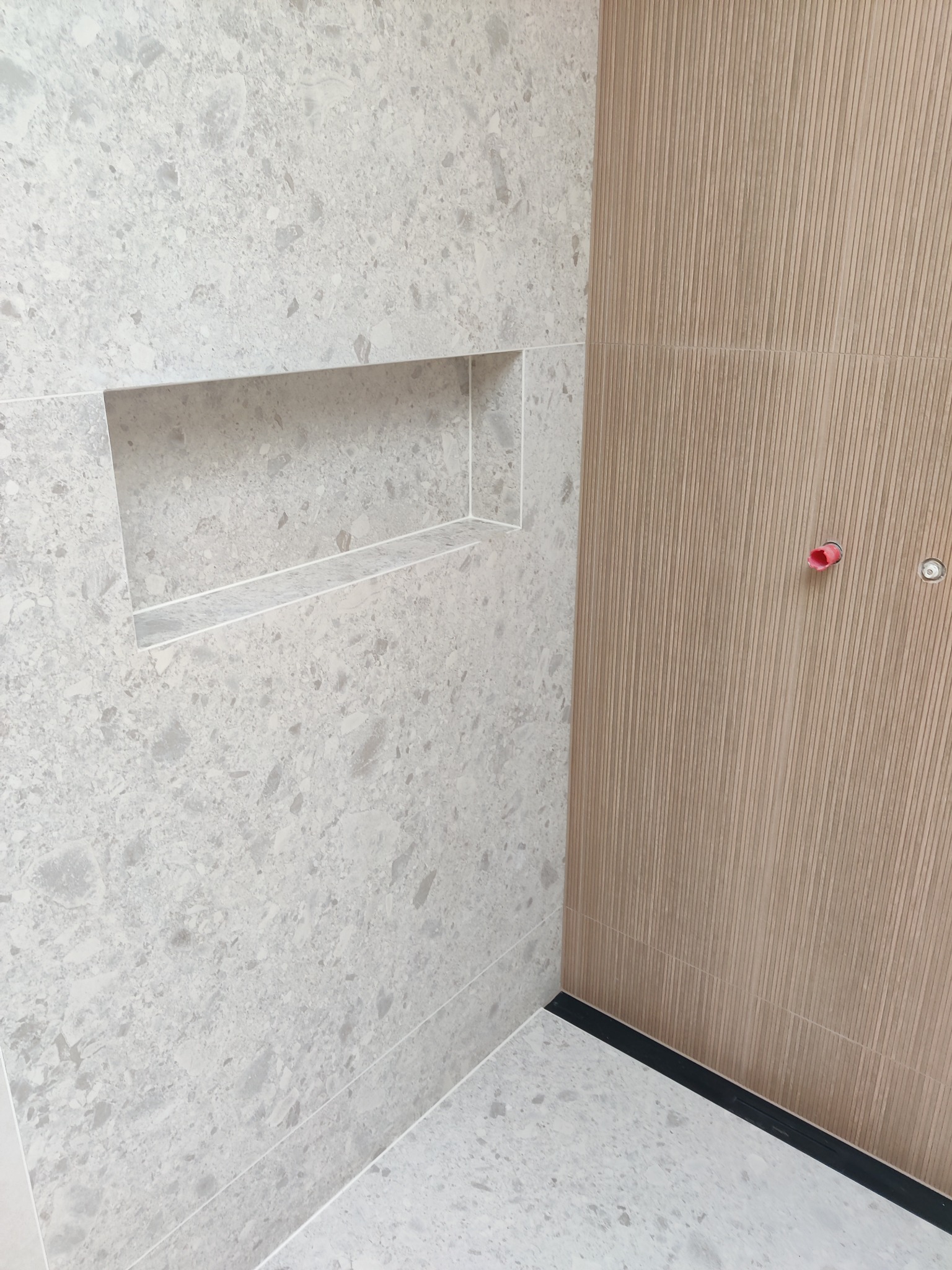 Wykończona łazienka z wnęką w ścianie wyłożonej szarymi płytkami lastryko i ścianą z płytek imitujących drewniane lamele. Widoczny fragment podłogi i czarny odpływ liniowy.