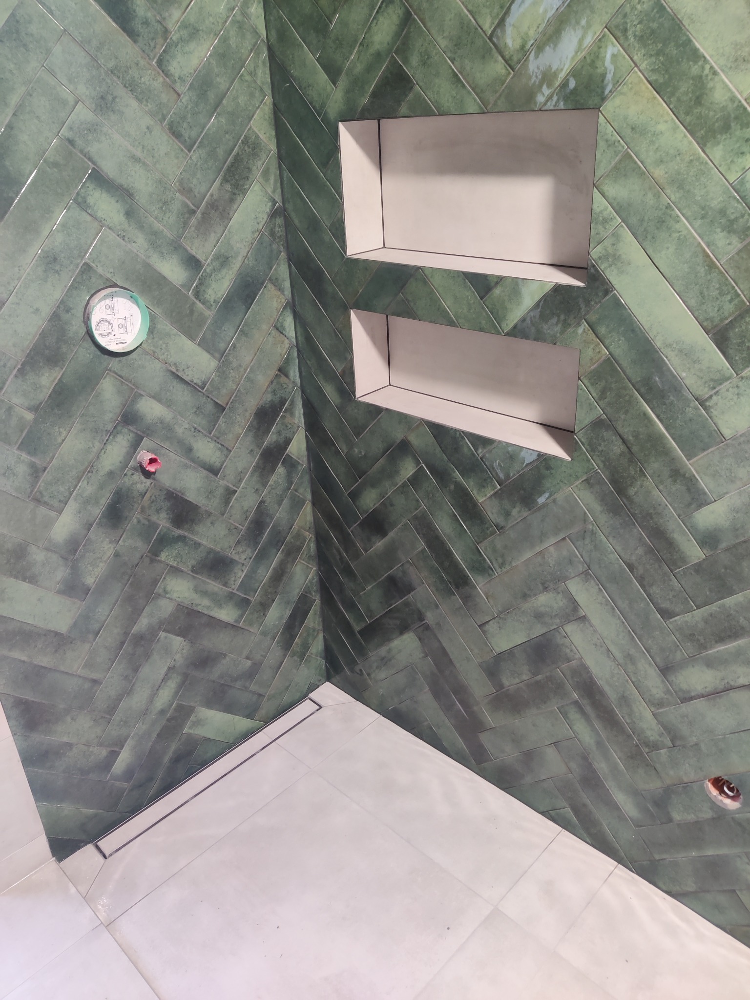 Ściana łazienki wyłożona zieloną, błyszczącą glazurą w jodełkę. Dwie wnęki w ścianie. Na podłodze jasne płytki. Widoczne elementy instalacji.