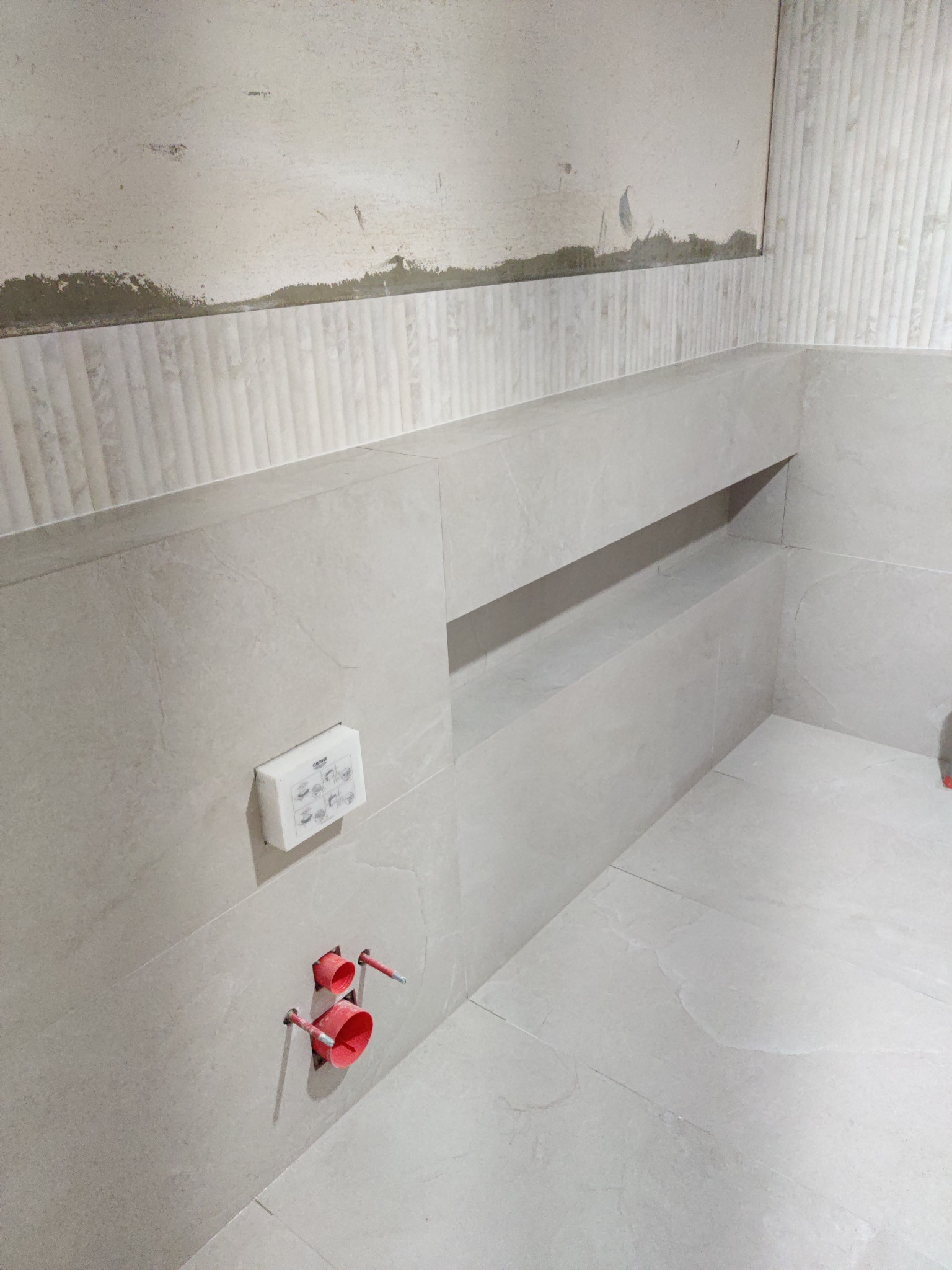 Jasne płytki imitujące kamień, częściowo ułożone w łazience z wnęką i wystającymi rurami. Widoczna puszka elektryczna i surowy fragment ściany.