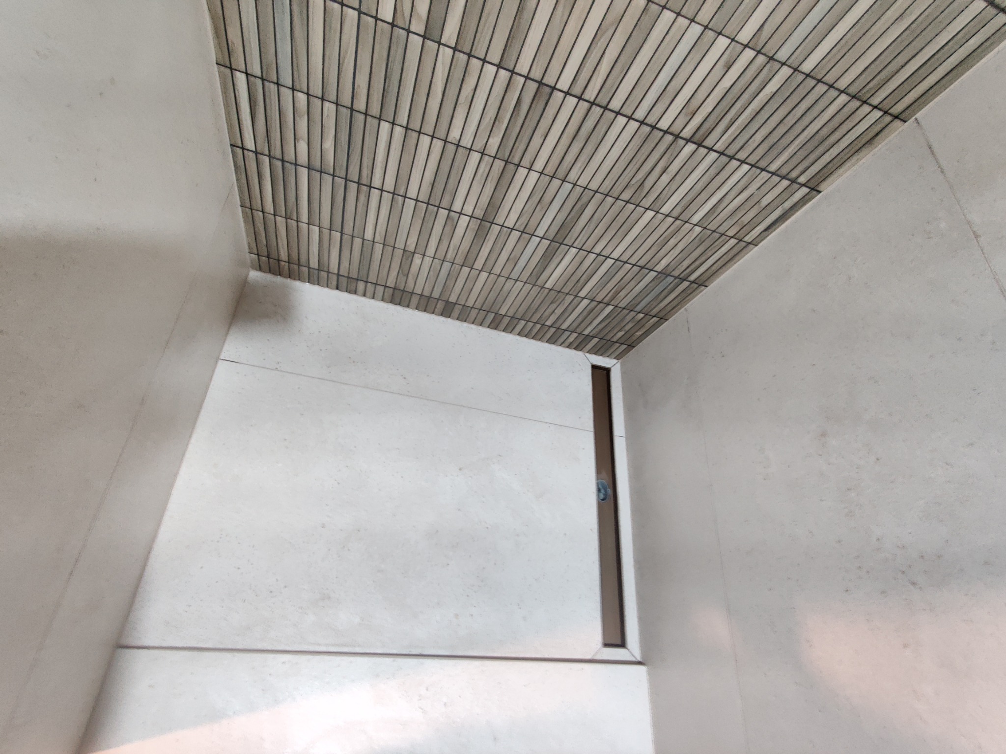 Wnętrze łazienki z wykończoną podłogą i ścianami w jasnych płytkach. Na suficie ułożone wąskie, poziome płytki imitujące drewno. Widoczny odpływ liniowy.