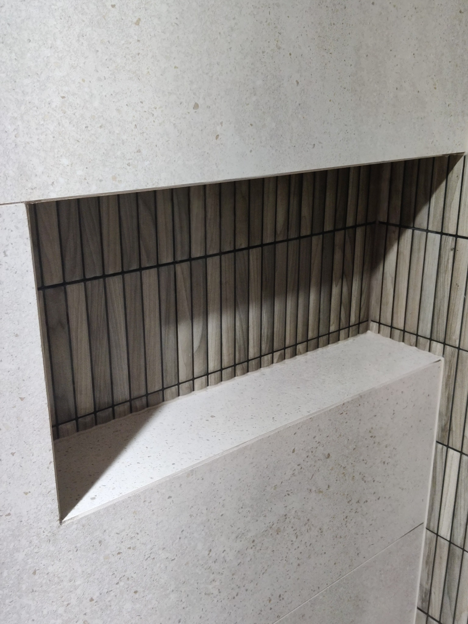 Wykończona wnęka w łazience z płytek imitujących kamień, z półką i tłem z płytek drewnopodobnych ułożonych pionowo.