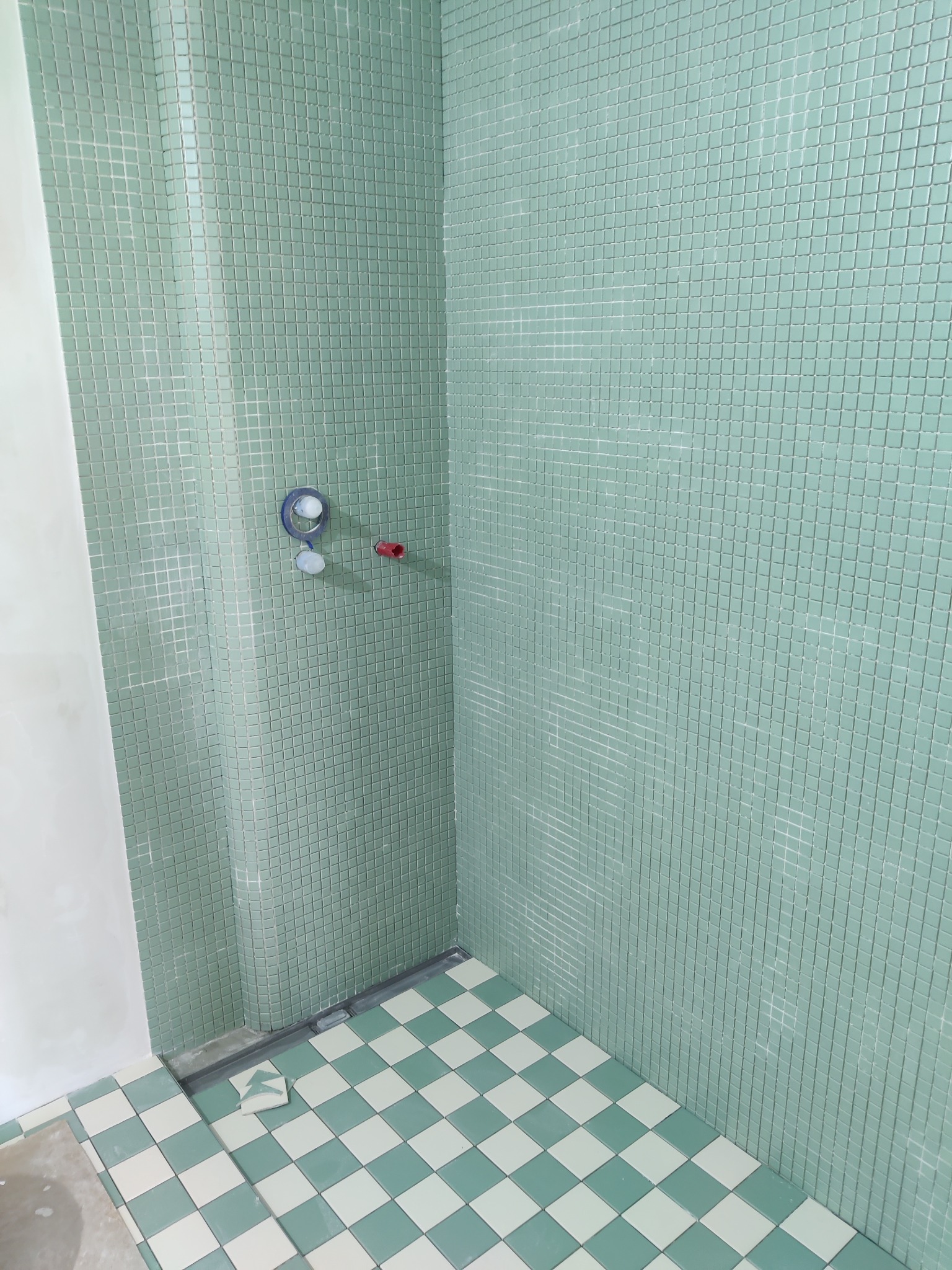 Ściana i podłoga w kabinie prysznicowej wyłożone mozaiką w odcieniach zieleni i bieli. Widoczne przyłącza wodne i odpływ liniowy. Częściowo położone płytki podłogowe.