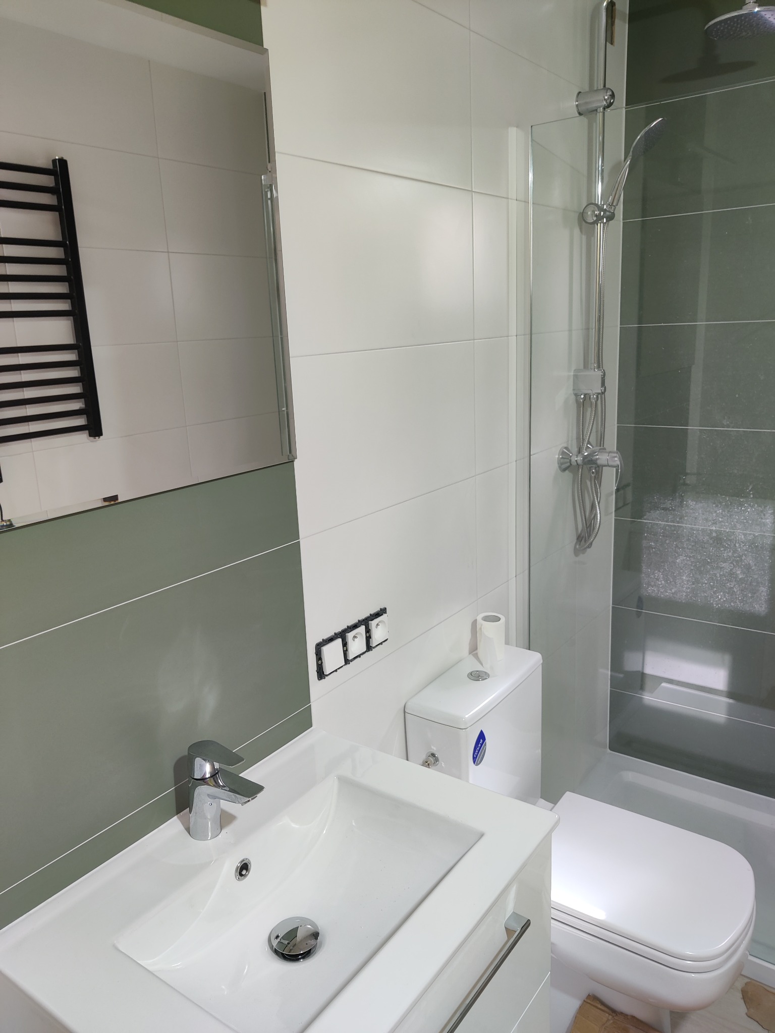 Nowoczesna łazienka po remoncie: białe płytki, szklana kabina prysznicowa, umywalka z szafką i grzejnik drabinkowy. Widoczne detale wykończenia i armatura.