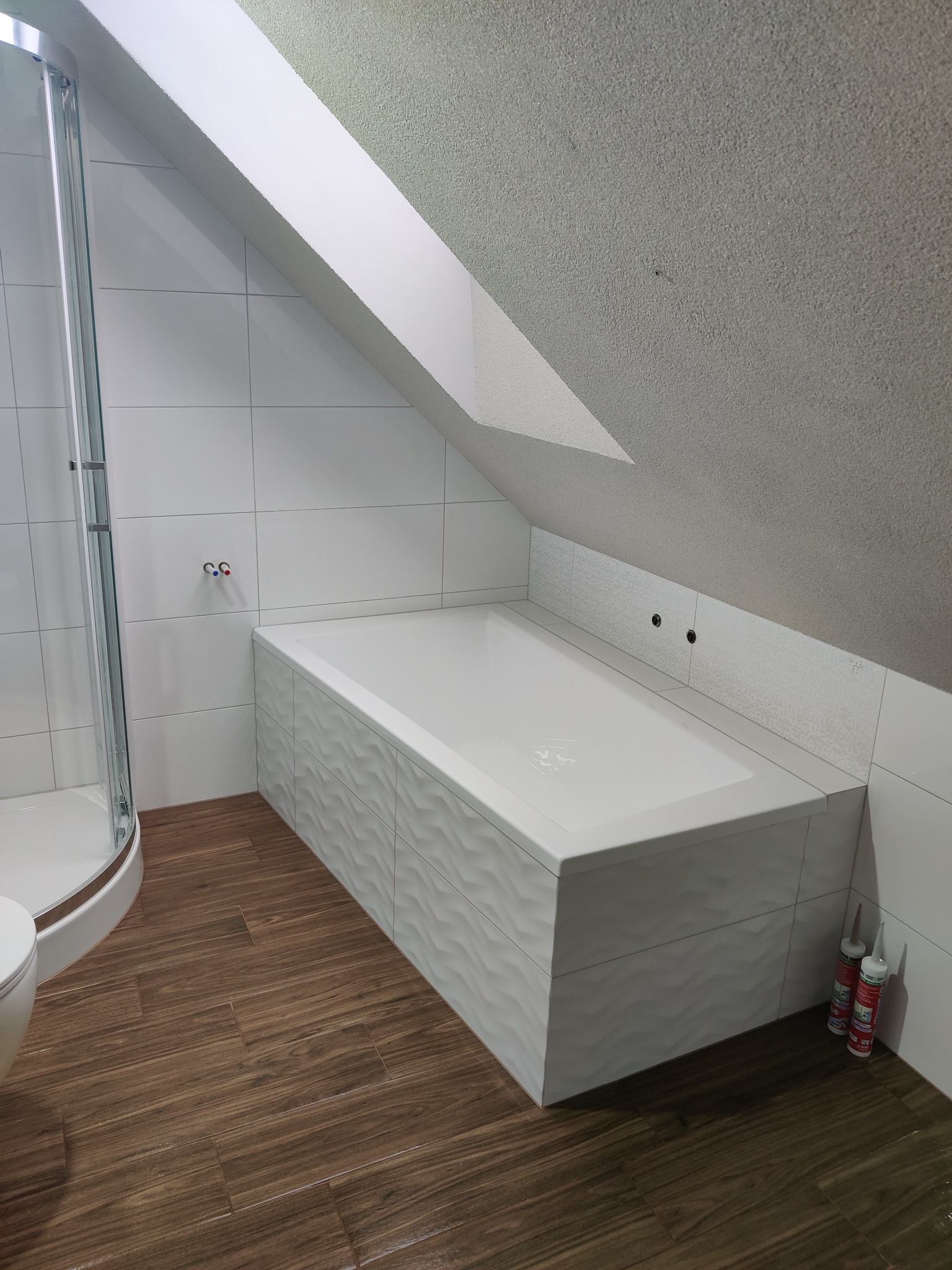 Biała wanna z dekoracyjnymi panelami w wykończonej łazience na poddaszu, widok na kabinę prysznicową i drewnopodobne płytki podłogowe. Dwa silikony przy wannie.