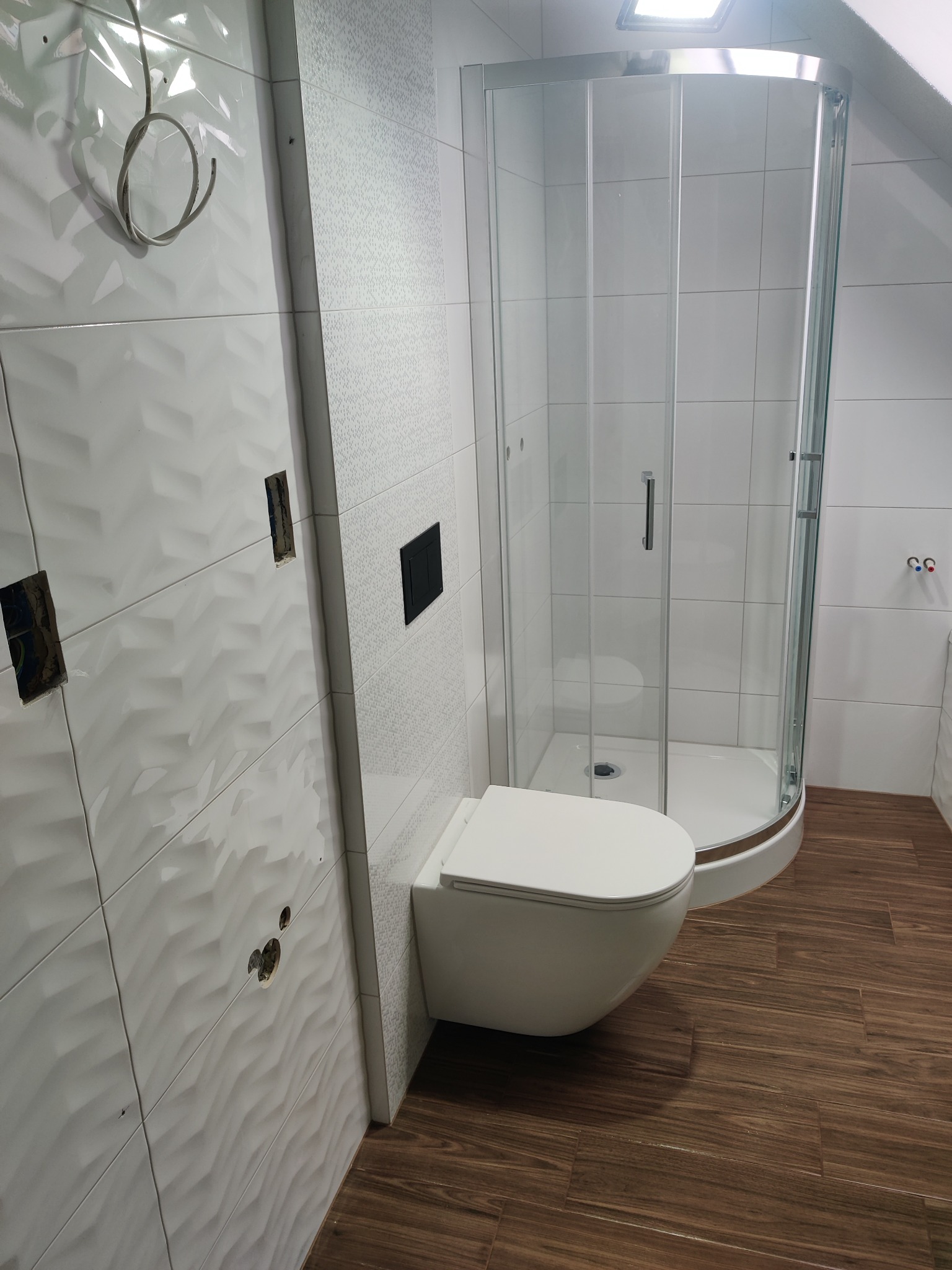 Nowoczesna łazienka z kabiną prysznicową i podwieszaną toaletą. Białe płytki 3D na ścianach, brązowe płytki imitujące drewno na podłodze. Częściowo wykończone otwory na gniazdka.