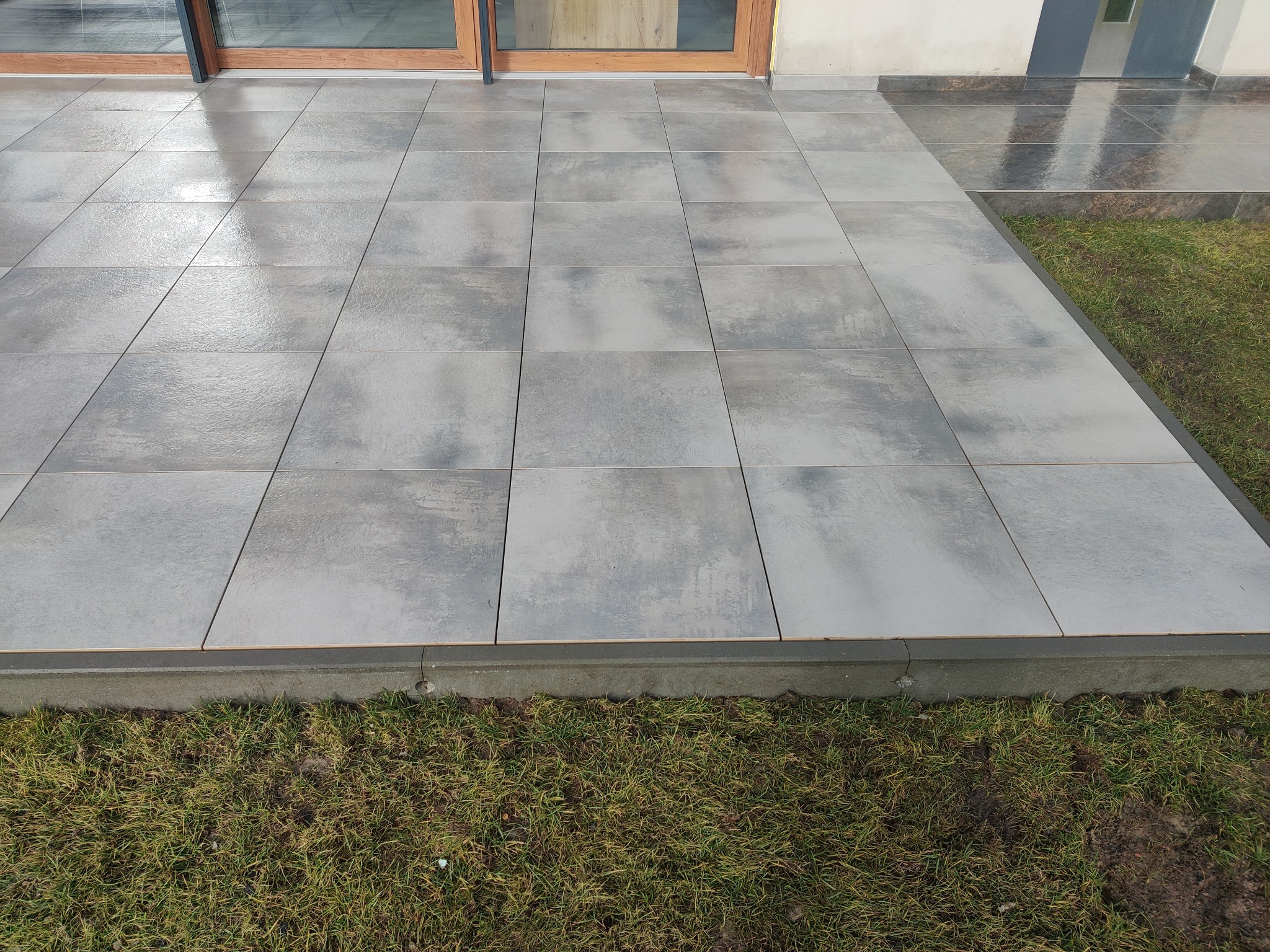 Szare, kwadratowe płytki tarasowe ułożone przy drzwiach balkonowych, z widocznym fragmentem trawnika. Minimalistyczny design, świeżo po deszczu, z połyskiem na powierzchni.