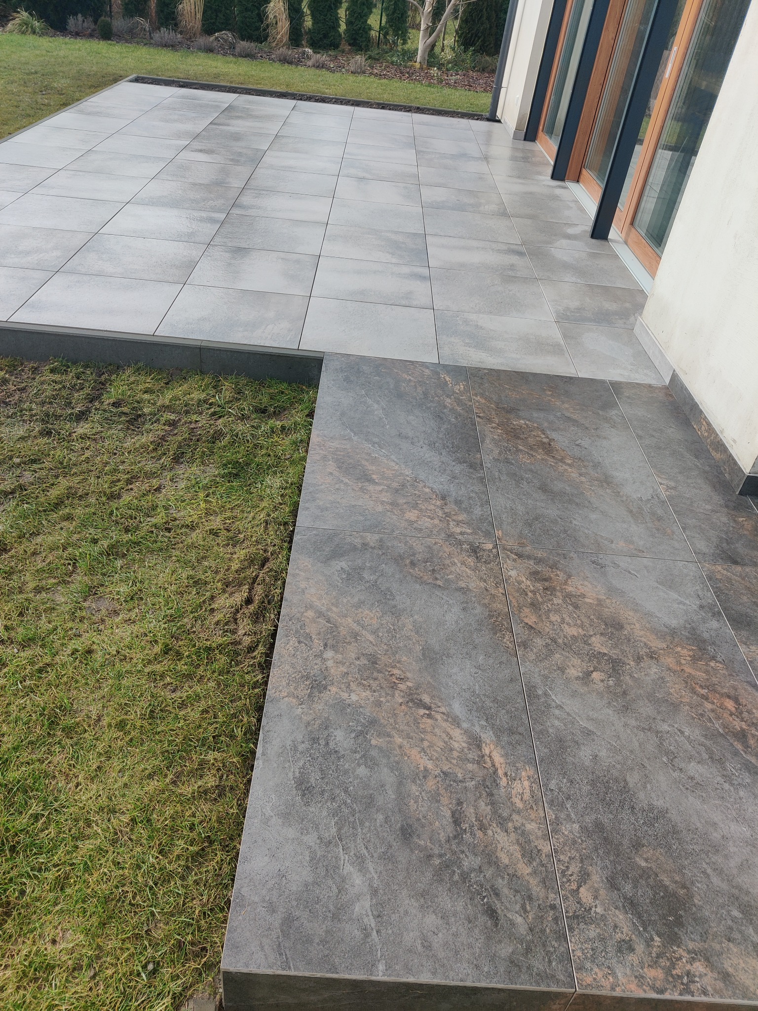 Ułożona, dwupoziomowa, szara, ceramiczna podłoga tarasu z ciemniejszymi płytkami na niższym poziomie, przylegająca do trawnika i budynku z oknami.