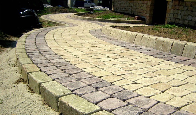 Kręta ścieżka z dwukolorowej kostki brukowej ułożona w ogrodzie z kamiennym budynkiem w tle.