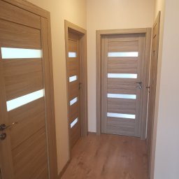 Aleksandr Balla - Korytarz z trzema drewnianymi drzwiami z poziomymi, mlecznymi przeszkleniami i panelami podłogowymi w odcieniach brązu.