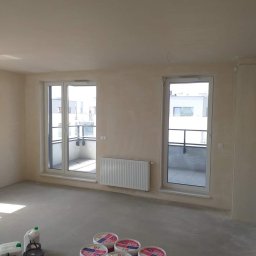 Remont4you - Przygotowane do malowania wnętrze: widoczne puszki z farbą Rapid, folie malarskie i narzędzia na podłodze, ściany po gruntowaniu, okna balkonowe z widokiem na sąsiedni budynek.