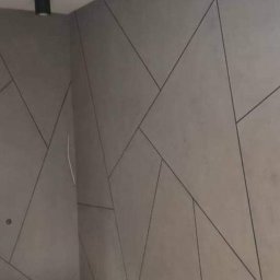 Remont4you - Ściana w szarym odcieniu z geometrycznym wzorem z czarnych linii, widoczne oświetlenie sufitowe i fragment sąsiedniego pomieszczenia.