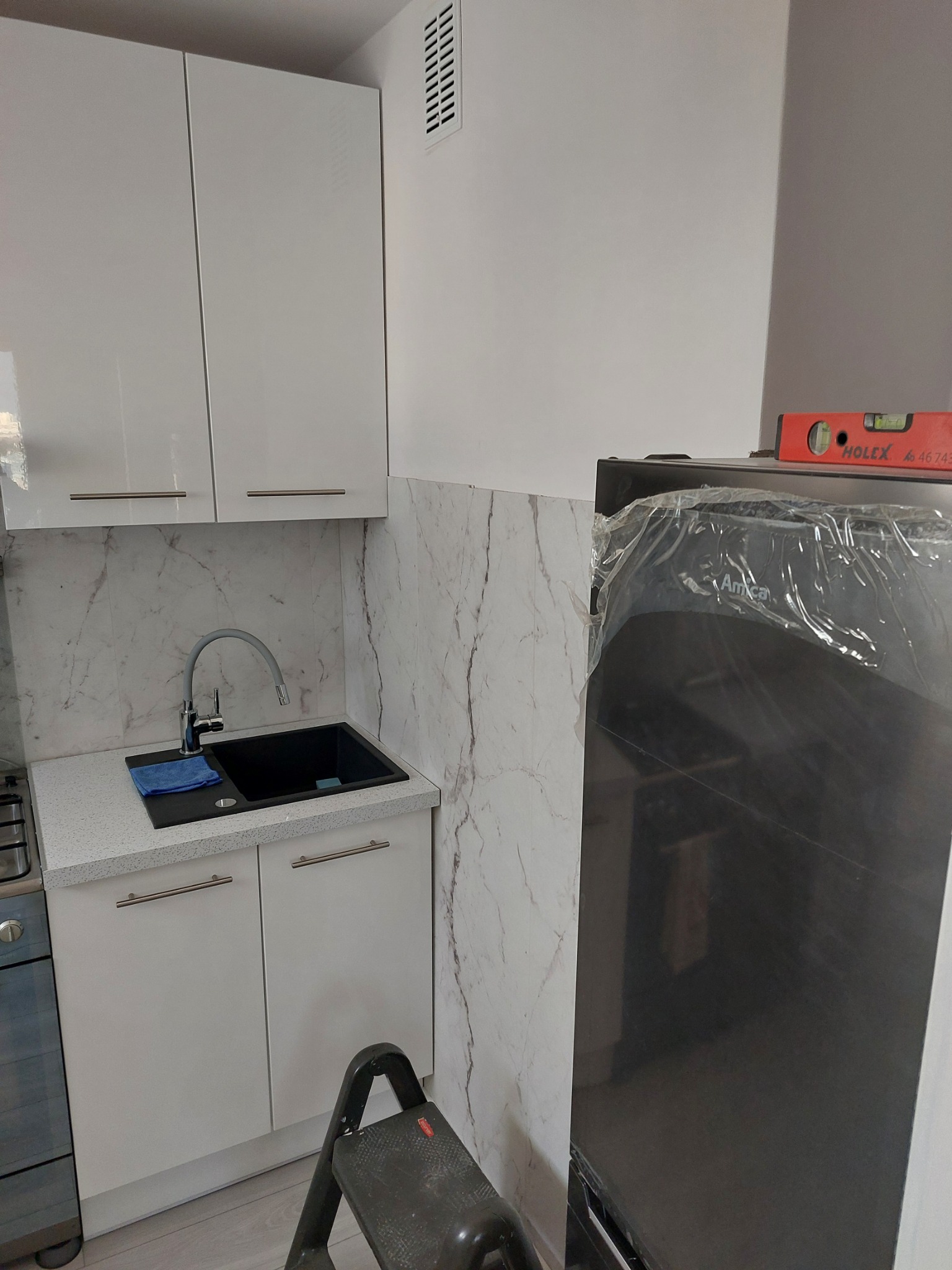 Fragment kuchni w trakcie montażu: białe szafki wiszące i dolne z czarnym zlewem, marmurowe panele ścienne, drabinka, lodówka Amica w folii ochronnej z poziomicą na górze.