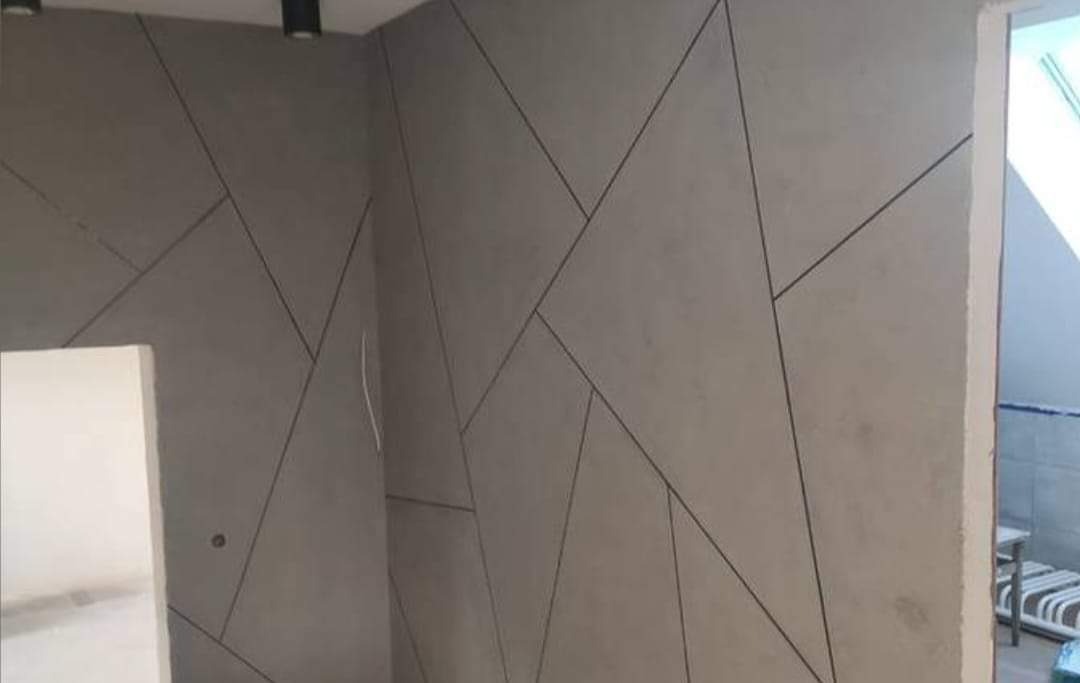 Ściana w szarym odcieniu z geometrycznym wzorem z czarnych linii, widoczne oświetlenie sufitowe i fragment sąsiedniego pomieszczenia.