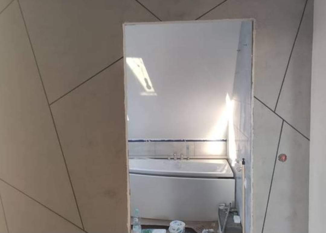 Fragment łazienki w trakcie wykańczania: ściana z geometrycznym wzorem w odcieniach szarości, widoczna przez otwór drzwiowy wanna i białe płytki. Częściowo wykończona ściana z widoczną instalacją...