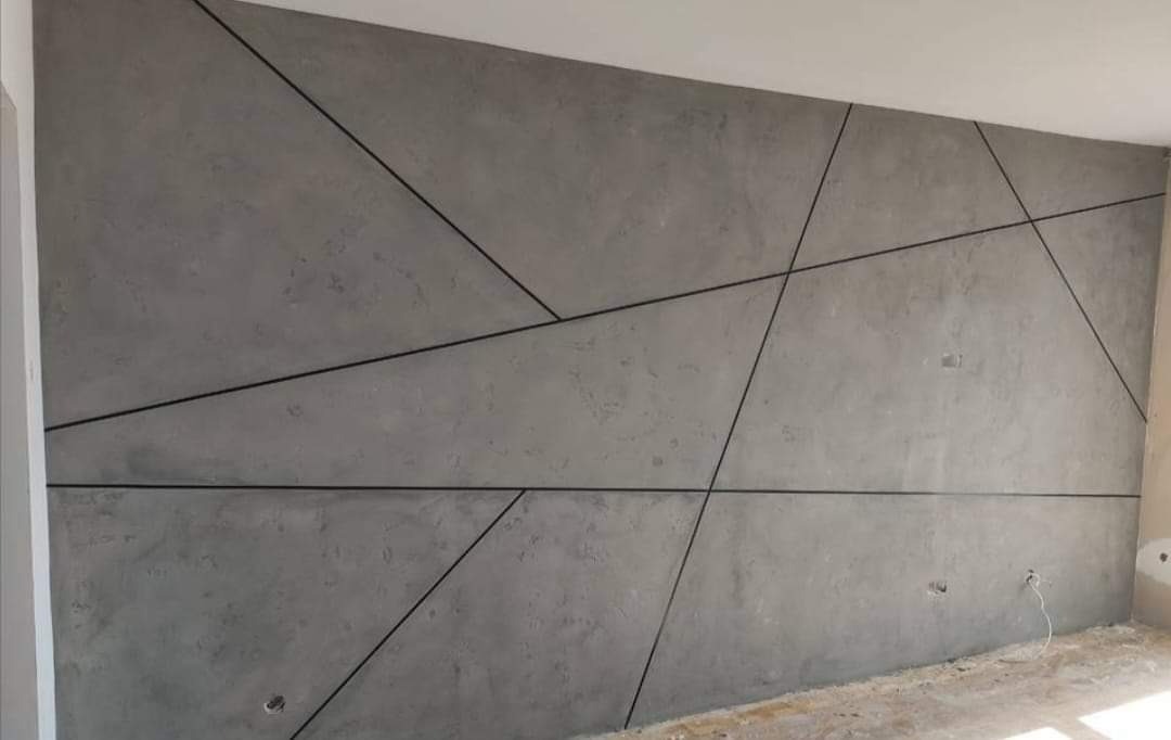 Betonowa ściana w odcieniach szarości z geometrycznym wzorem utworzonym przez czarne, cienkie listwy dekoracyjne.