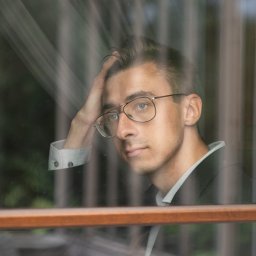 Krzysztof Sałaciński - Młody mężczyzna w okularach, oparty o szybę okna z drewnianą ramą, w marynarce i koszuli z mankietami, zamyślony wyraz twarzy.