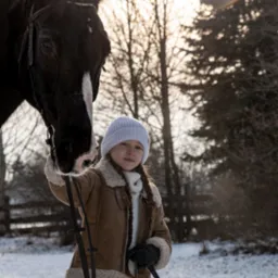 Dziewczynka w zimowej kurtce i czapce trzyma czarnego konia za uzdę na śniegu, z drzewami w tle.