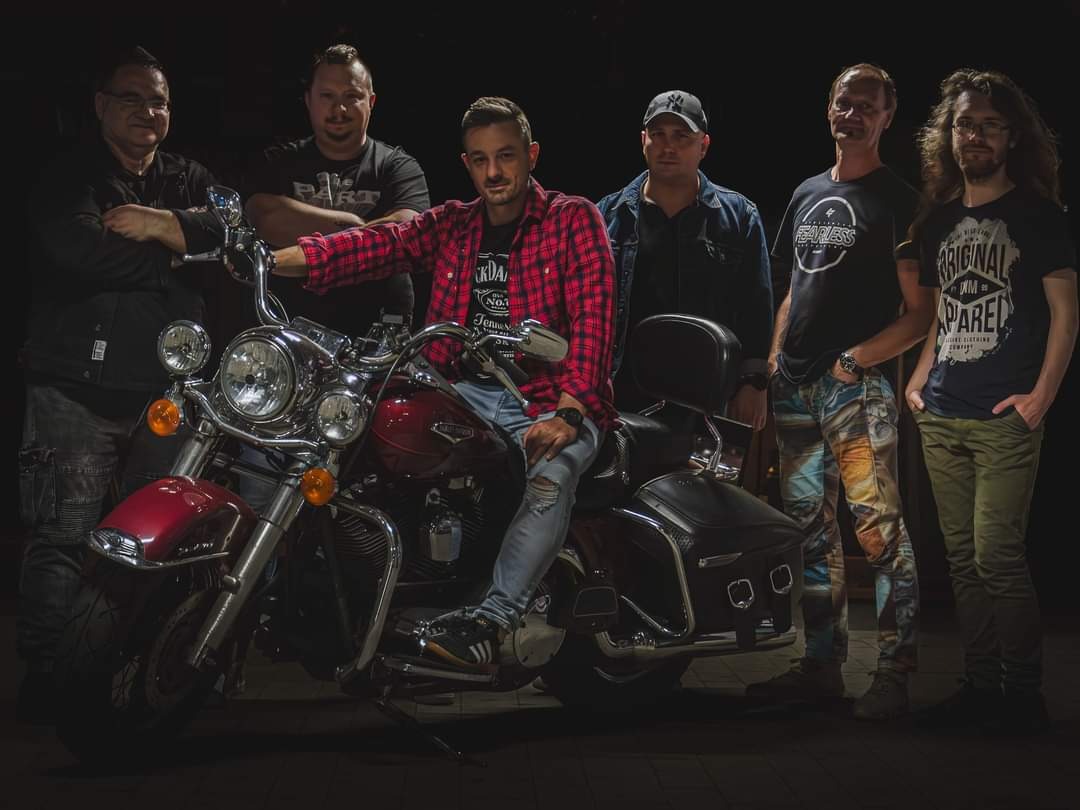 Zespół muzyczny pozuje z czerwonym motocyklem typu cruiser w ciemnym otoczeniu, lider w czerwonej koszuli w kratę siedzi na motocyklu.