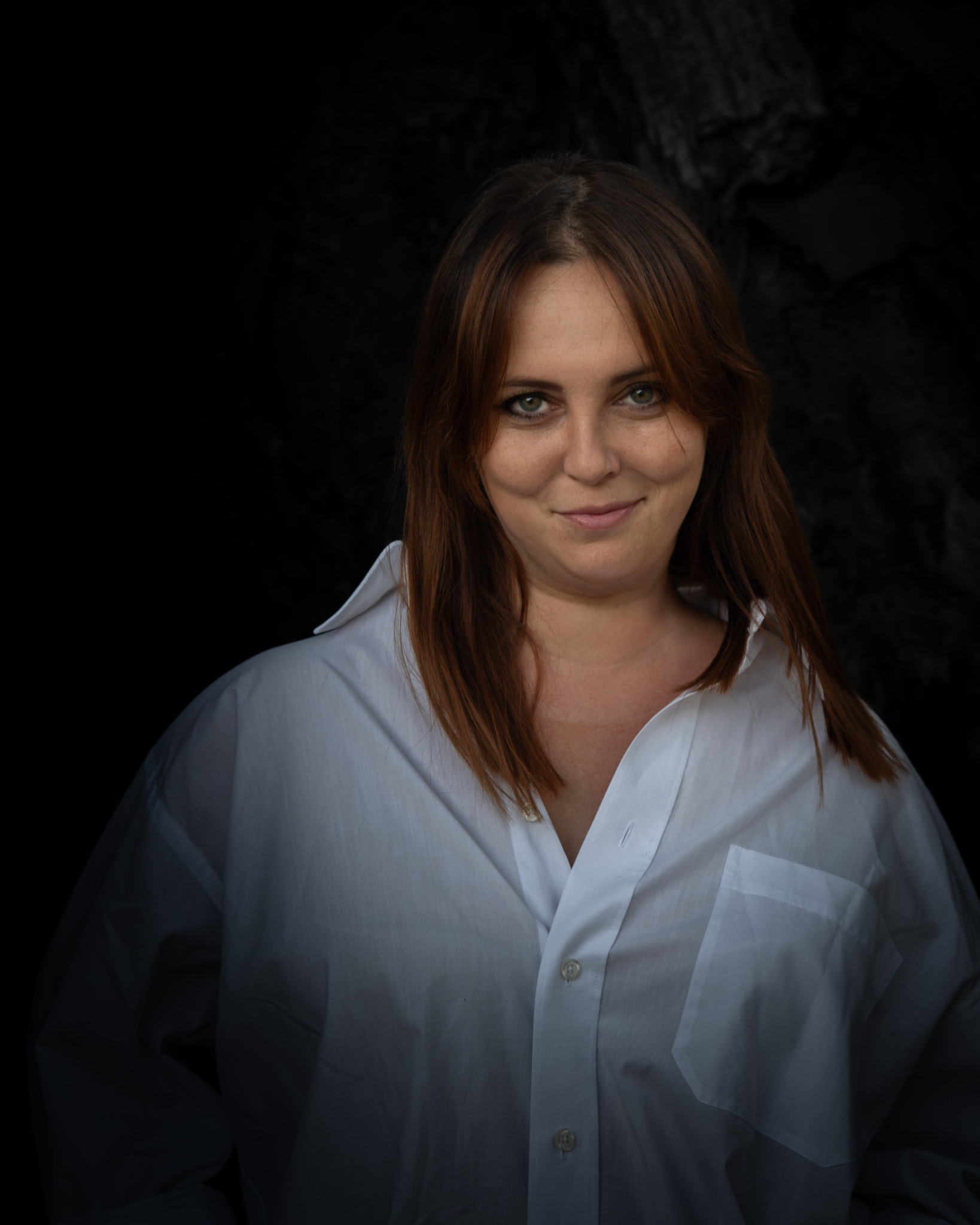 Portret kobiety o zielonych oczach i kasztanowych włosach, ubranej w białą koszulę na tle ciemnego, teksturalnego tła.