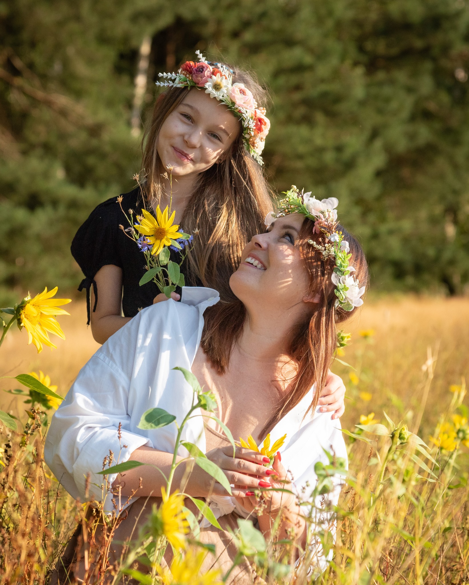 Matka i córka w wiankach z kwiatów na głowach, na tle pola słoneczników i lasu, córka trzyma bukiecik polnych kwiatów.