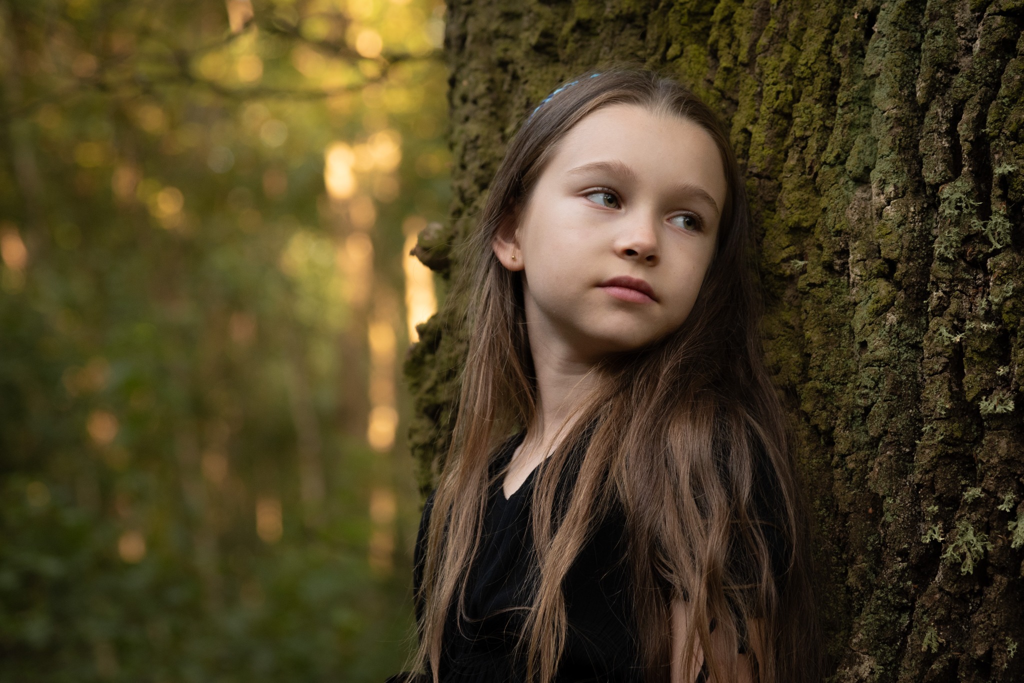 Młoda dziewczyna z długimi brązowymi włosami opiera się o omszałe drzewo, zamyślona, w tle rozmyty leśny krajobraz z promieniami słońca.