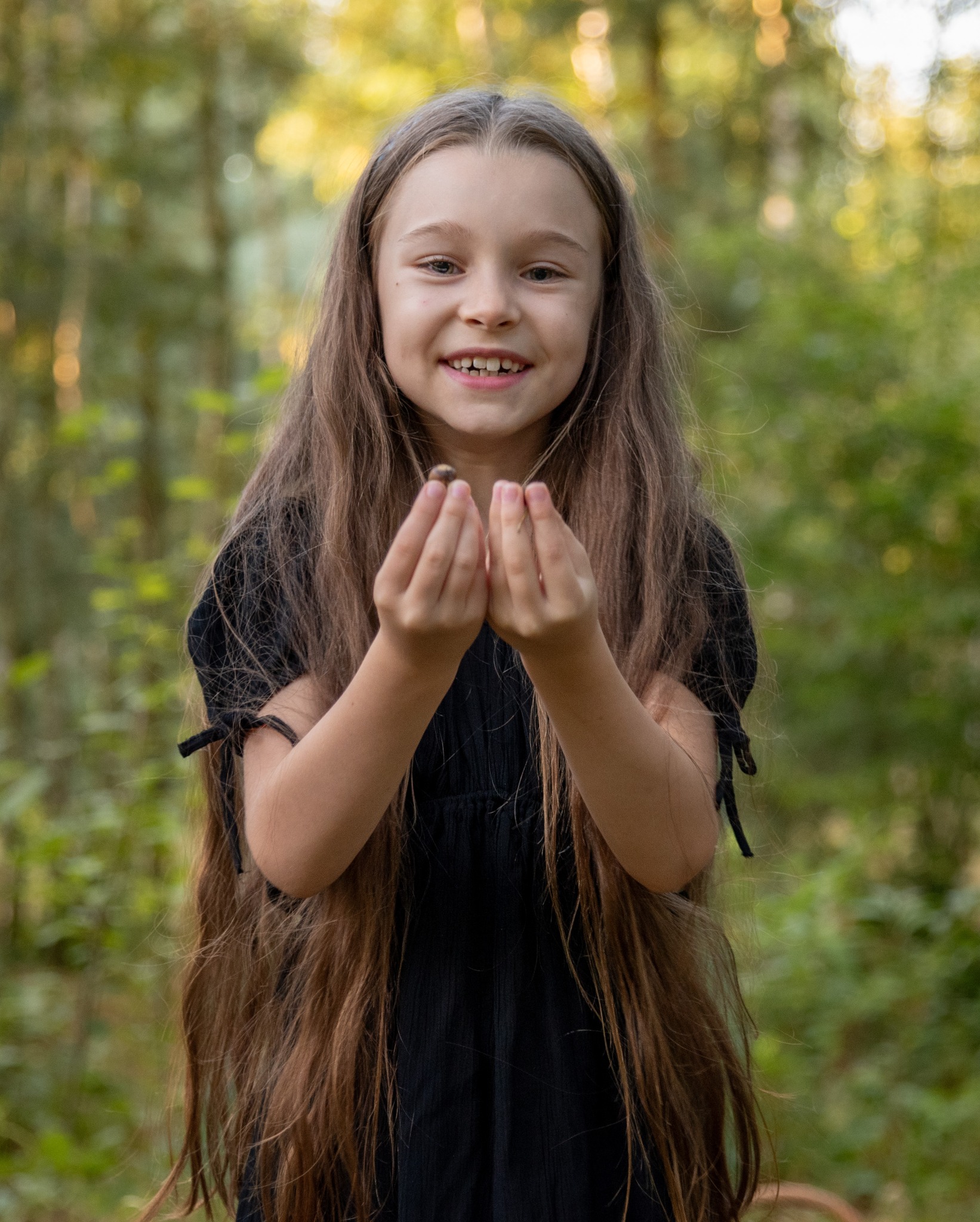 Uśmiechnięta dziewczynka w czarnej sukience trzymająca w dłoniach żołądź na tle zamazanego, zielonego lasu.