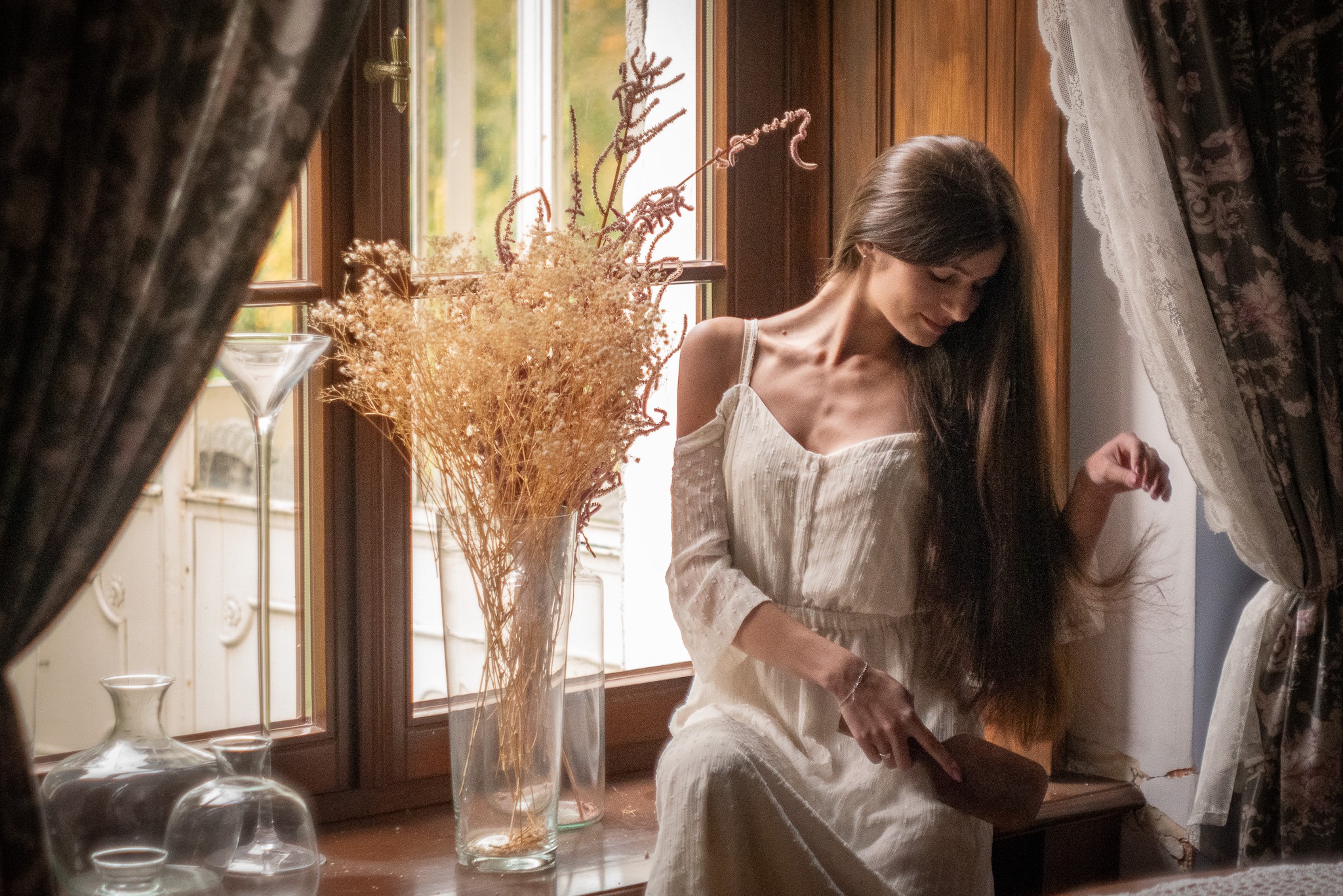 Kobieta w białej sukience siedzi na parapecie okna, z bukietem suszonych kwiatów w wazonie i lustrem obok, w otoczeniu zasłon w stylu vintage.