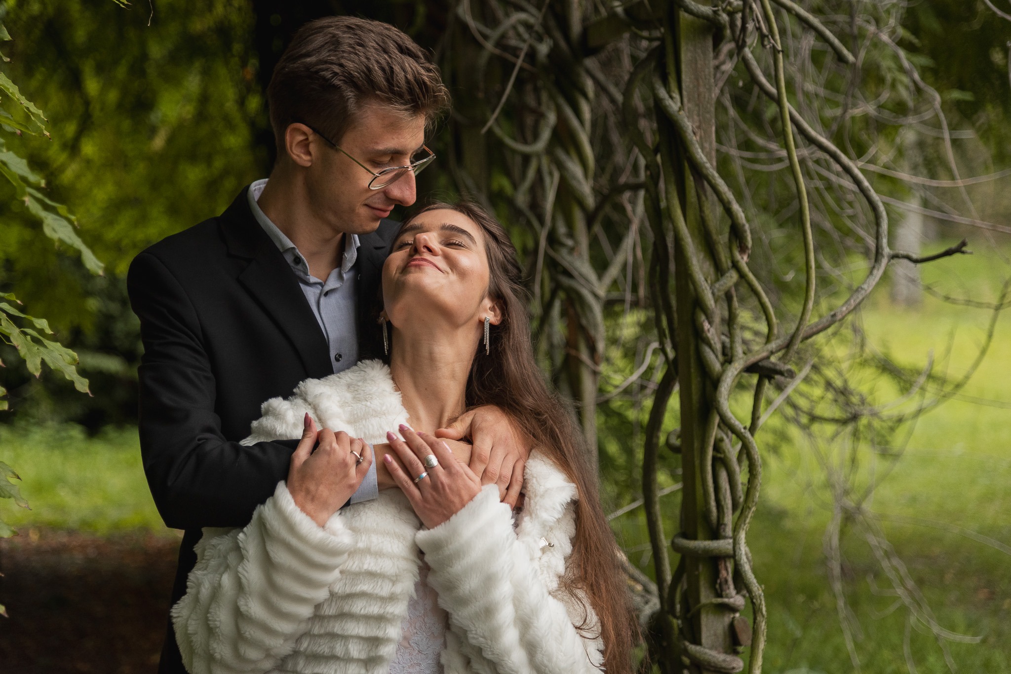 Uśmiechnięta para w objęciach na tle drewnianej konstrukcji porośniętej pnączami, kobieta w białym futerku i z błyszczącymi kolczykami.