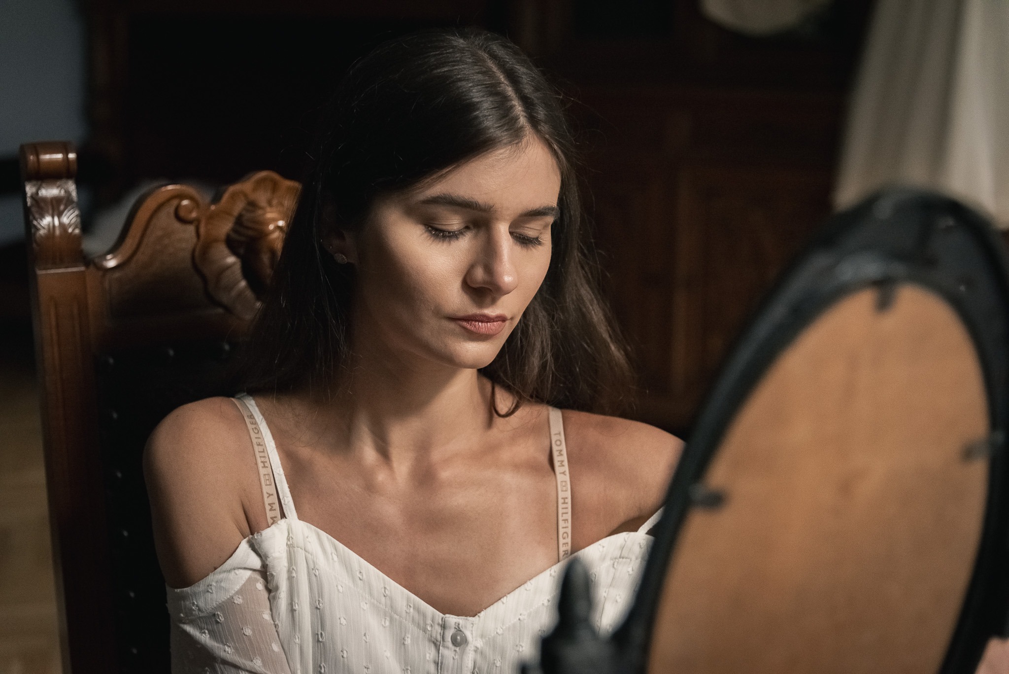 Młoda kobieta w białej bluzce z odkrytymi ramionami, siedząca na drewnianym krześle, spogląda w dół, częściowo zasłonięta przez owalne lustro z czarną ramą.