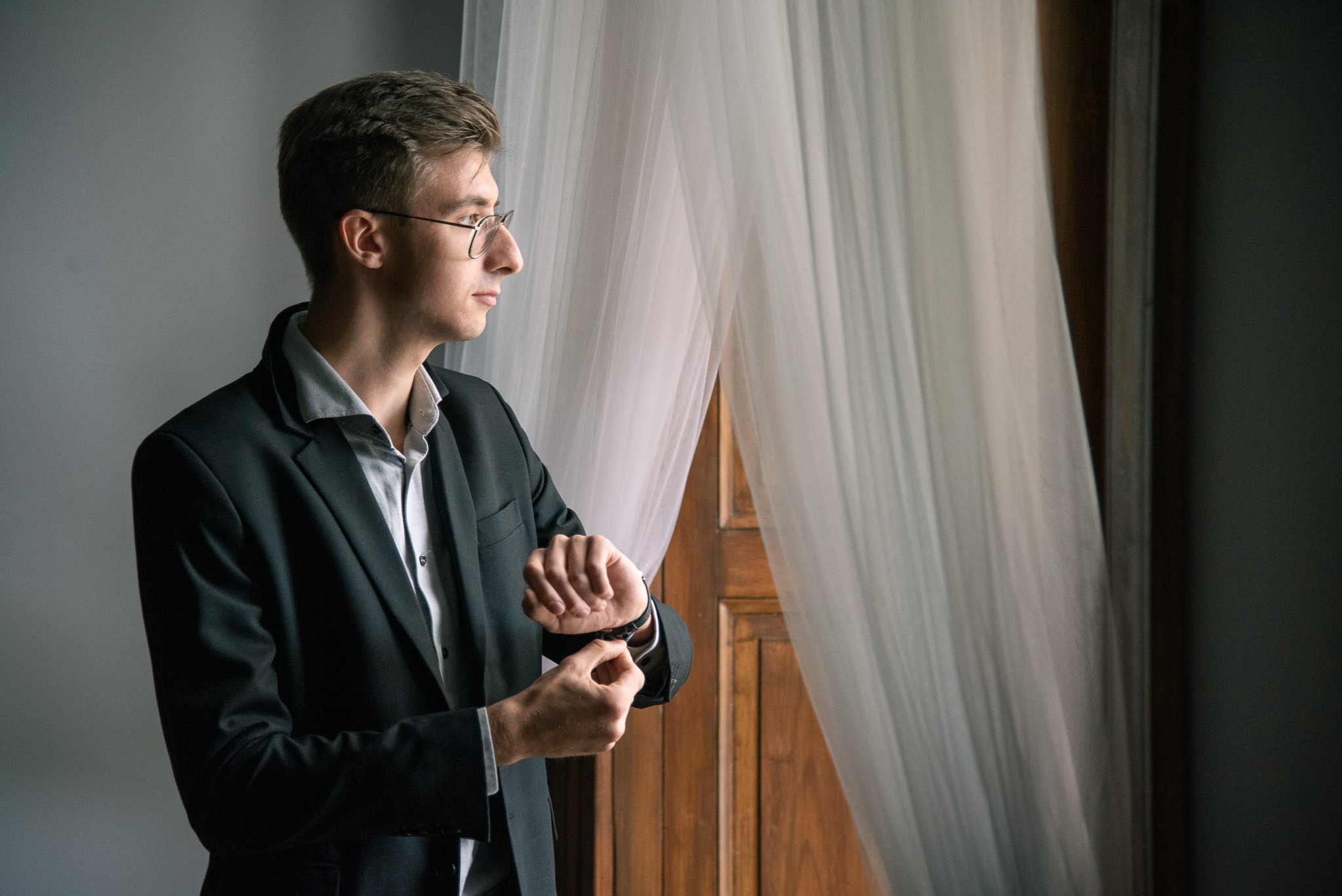 Młody mężczyzna w garniturze, stojący przy oknie z białymi zasłonami i sprawdzający godzinę na zegarku, profil.