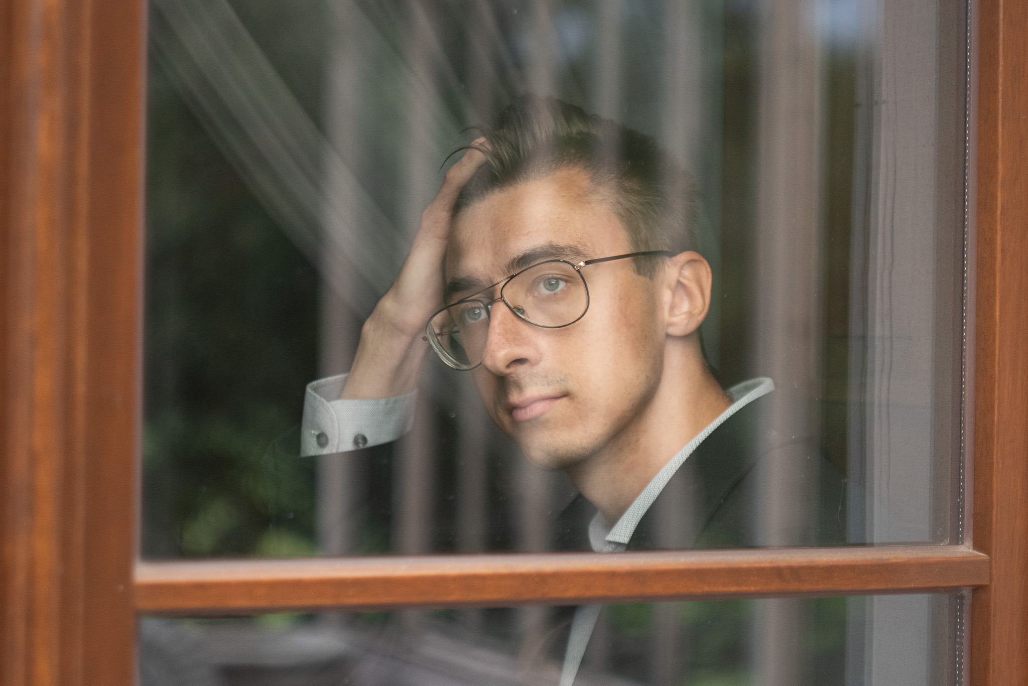 Młody mężczyzna w okularach, oparty o szybę okna z drewnianą ramą, w marynarce i koszuli z mankietami, zamyślony wyraz twarzy.
