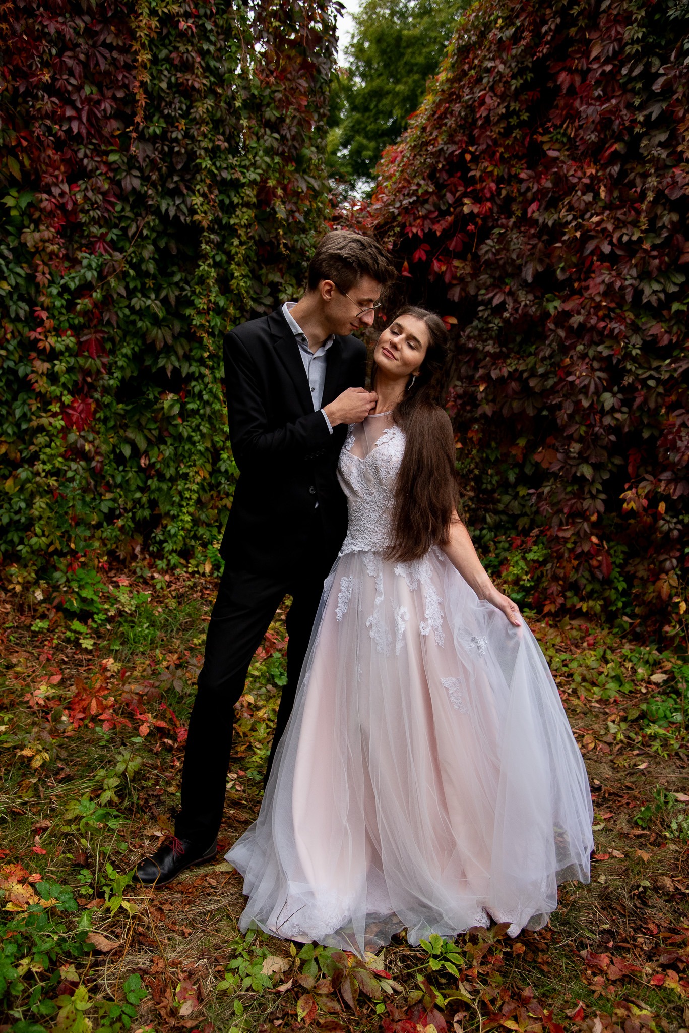 Para młoda w plenerze: pan młody w czarnym garniturze delikatnie dotyka szyi panny młodej w białej sukni z koronkowymi zdobieniami na tle ściany z czerwonych liści.