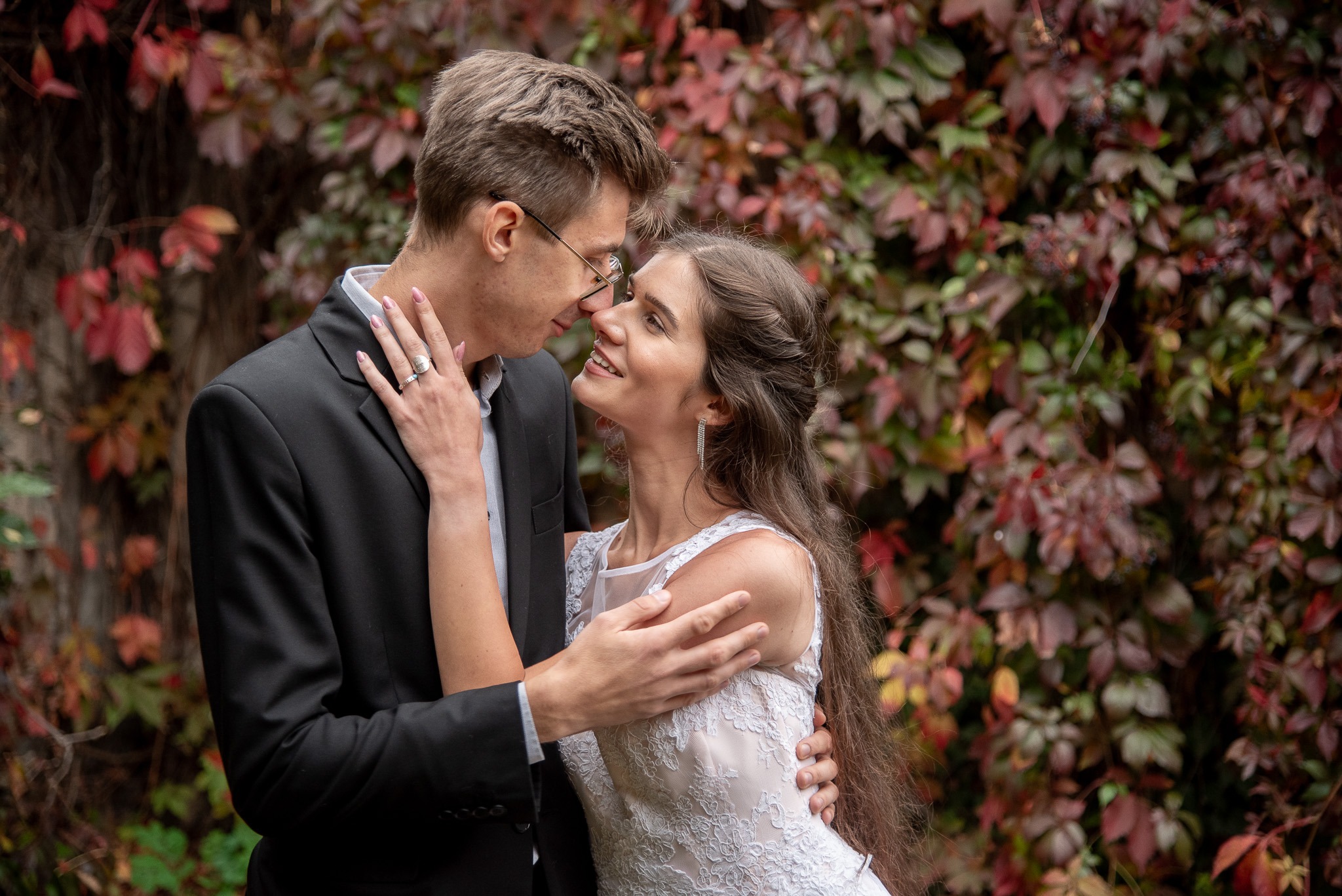 Uśmiechnięta panna młoda w koronkowej sukni, obejmowana przez pana młodego w czarnym garniturze na tle ściany z czerwonych i zielonych liści.