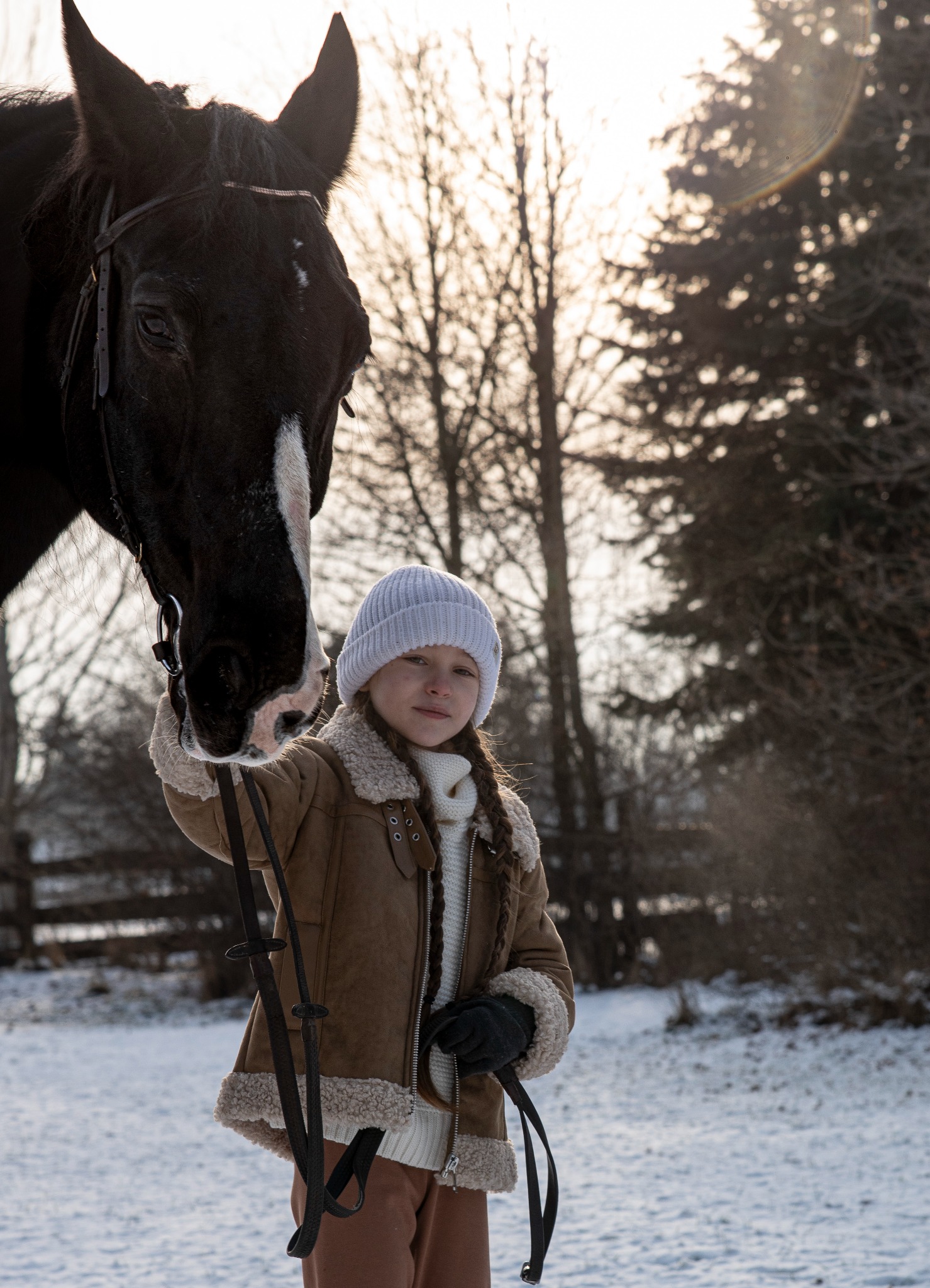Dziewczynka w zimowej kurtce i czapce trzyma czarnego konia za uzdę na śniegu, z drzewami w tle.