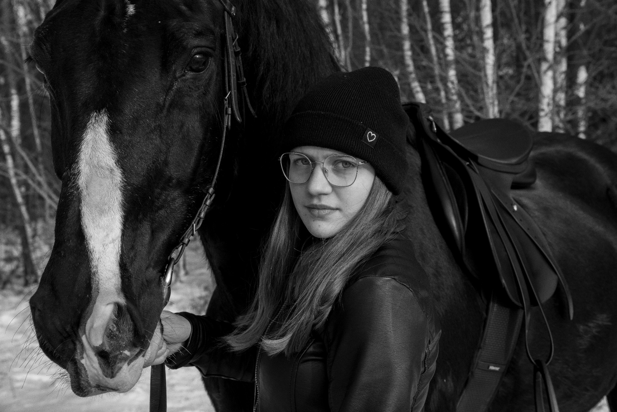 Czarno-białe zdjęcie portretowe młodej kobiety w okularach i czapce z serduszkiem, stojącej obok ciemnego konia z siodłem, na tle drzew.