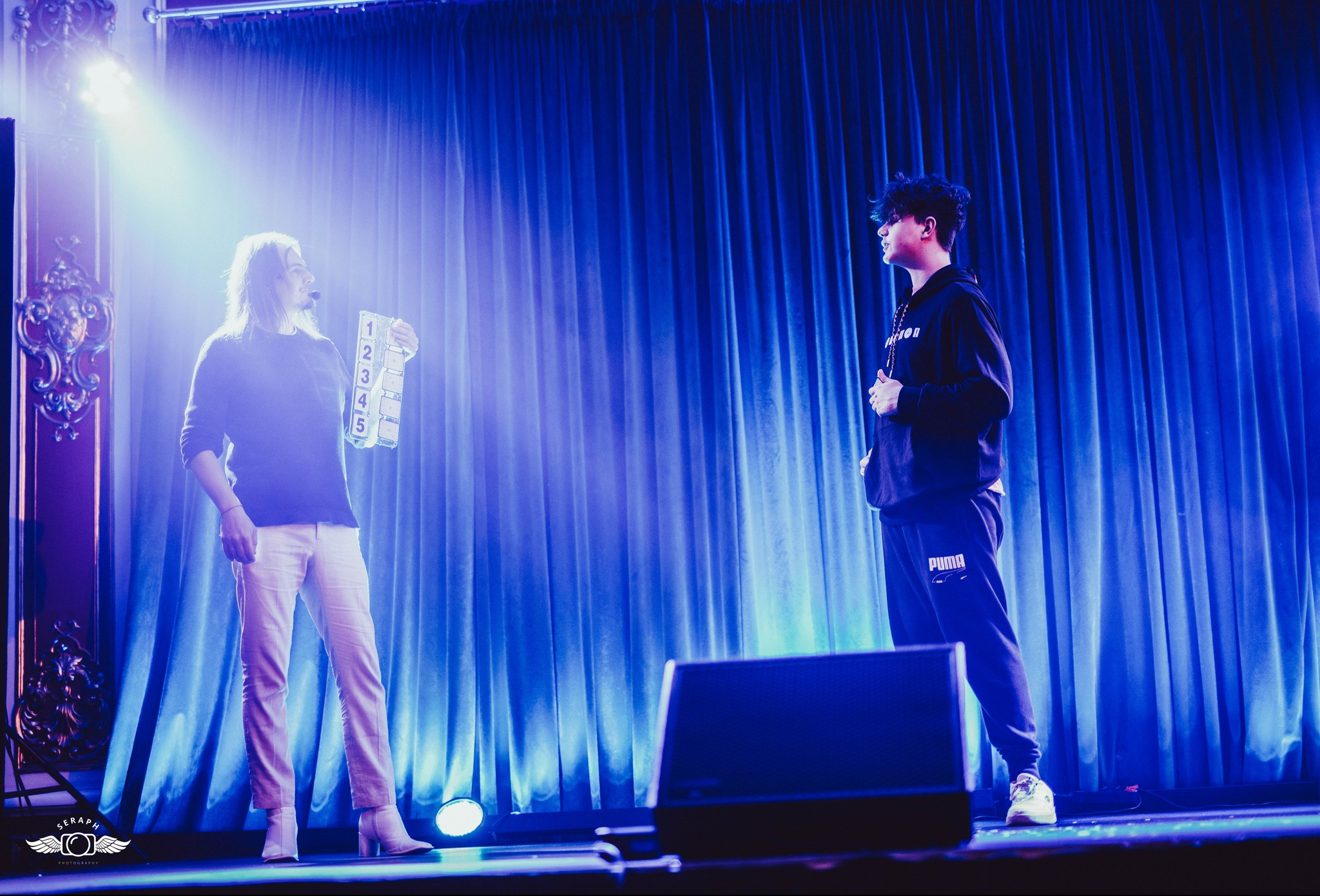 Iluzjonista w czarnym swetrze i jasnych spodniach trzyma rekwizyt z numerami, na scenie z niebieskimi zasłonami, obok stoi młody mężczyzna w dresie.