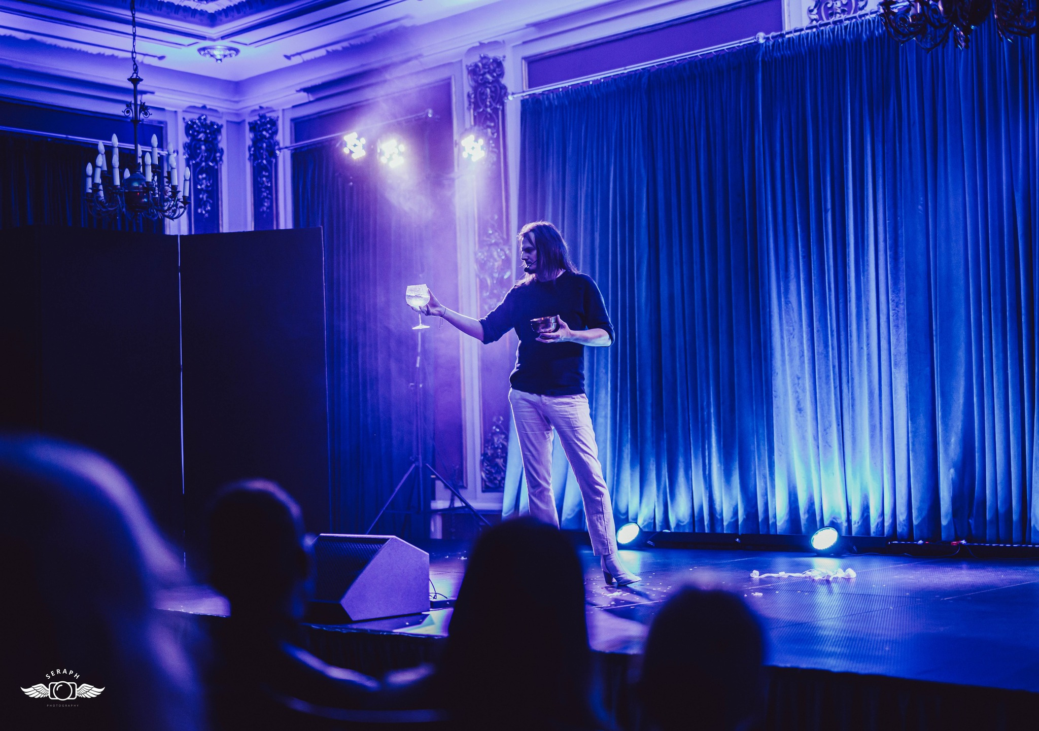 Iluzjonista w trakcie występu na scenie oświetlonej niebieskim światłem, trzymający kieliszek z płynem i małą miseczkę, w tle zasłony i dekoracje w stylu retro.