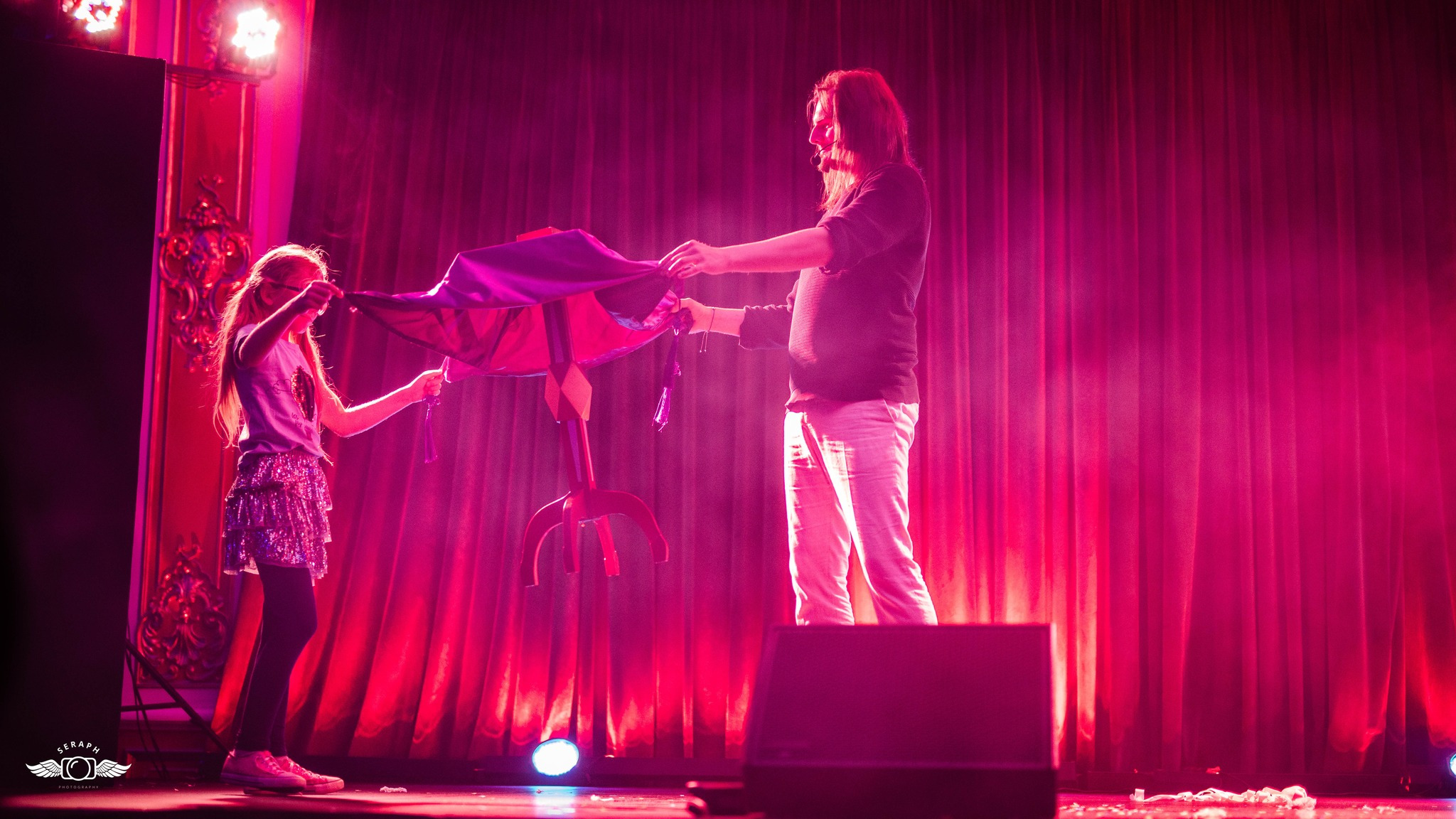 Iluzjonista z długimi włosami, w czarnej koszuli i jasnych spodniach, prezentuje sztuczkę z małą dziewczynką na scenie oświetlonej czerwonymi światłami, używając stolika z trzema nogami...