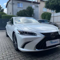 Biały samochód Lexus ES z tabliczką z nazwą firmy na tle kostki brukowej i budynku mieszkalnego.