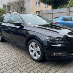 Pomydleni - Czarny samochód kombi marki Skoda Superb zaparkowany na brukowanej nawierzchni, z widocznym niebieskim samochodem w tle i budynkiem mieszkalnym.