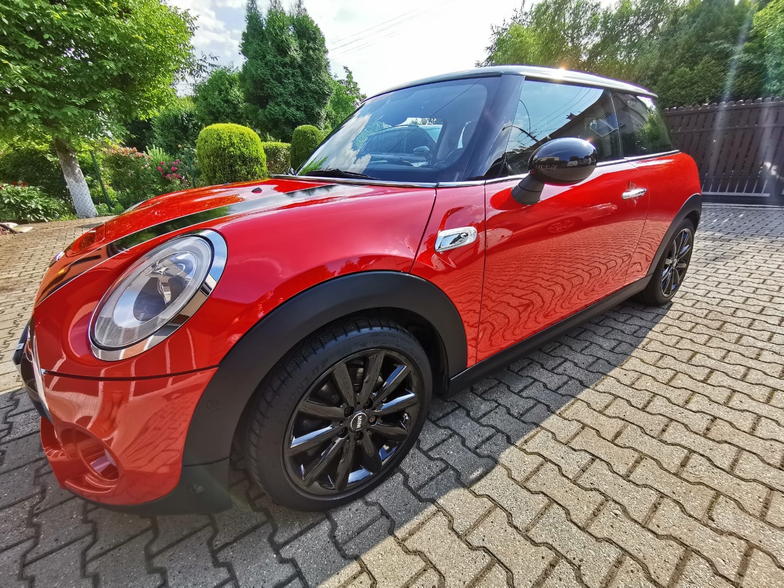 Czerwony Mini Cooper po umyciu, z widocznymi kroplami wody na lakierze, stojący na kostce brukowej w słoneczny dzień.