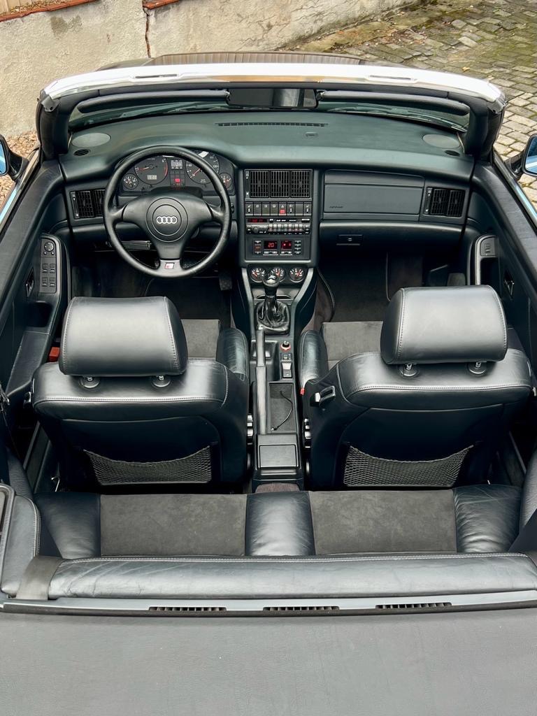 Wnętrze czarnego kabrioletu Audi z widoczną deską rozdzielczą, kierownicą z logo Audi, skórzanymi fotelami i manualną skrzynią biegów, sfotografowane z góry.