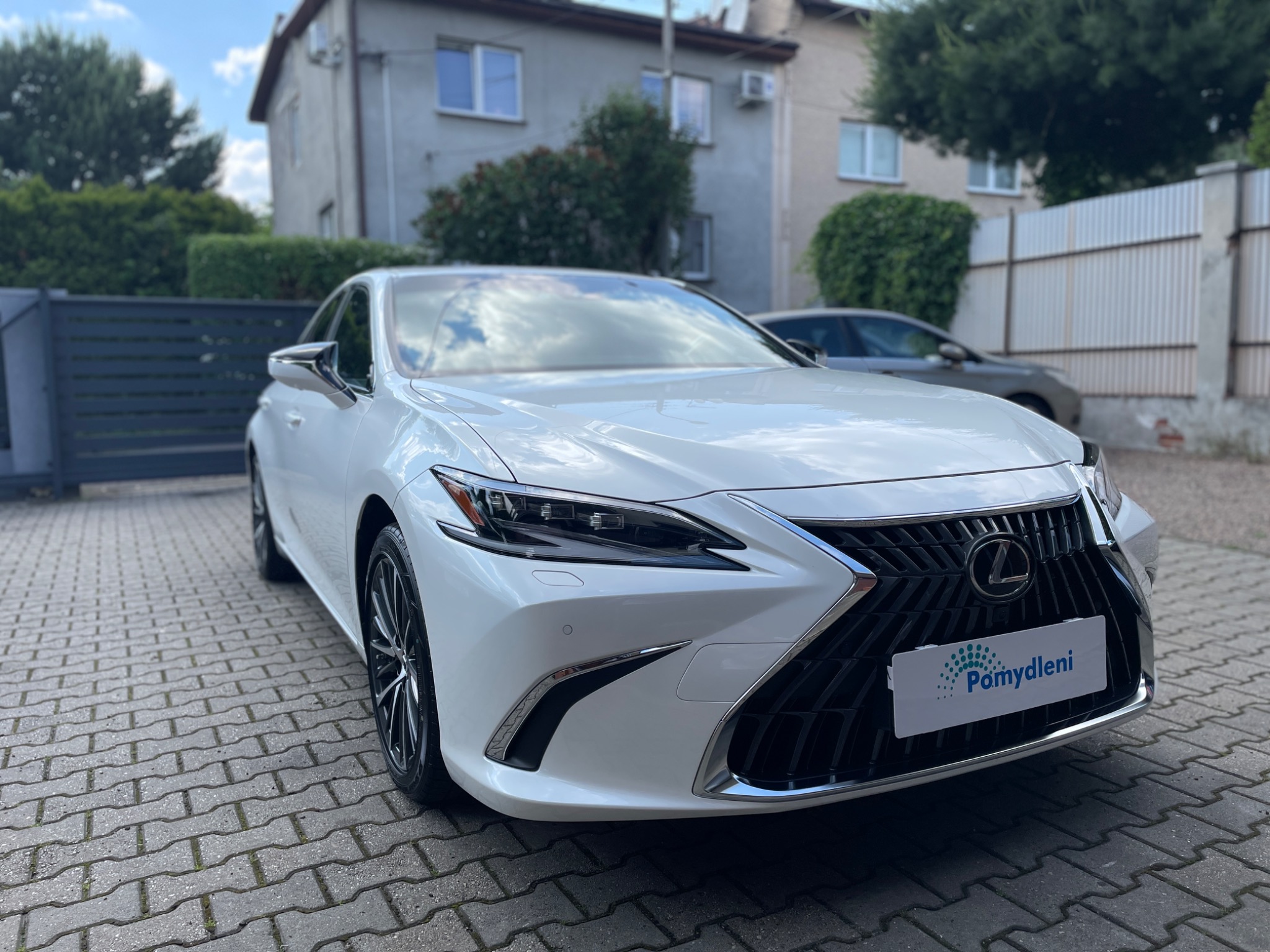 Biały samochód Lexus ES z tabliczką z nazwą firmy na tle kostki brukowej i budynku mieszkalnego.