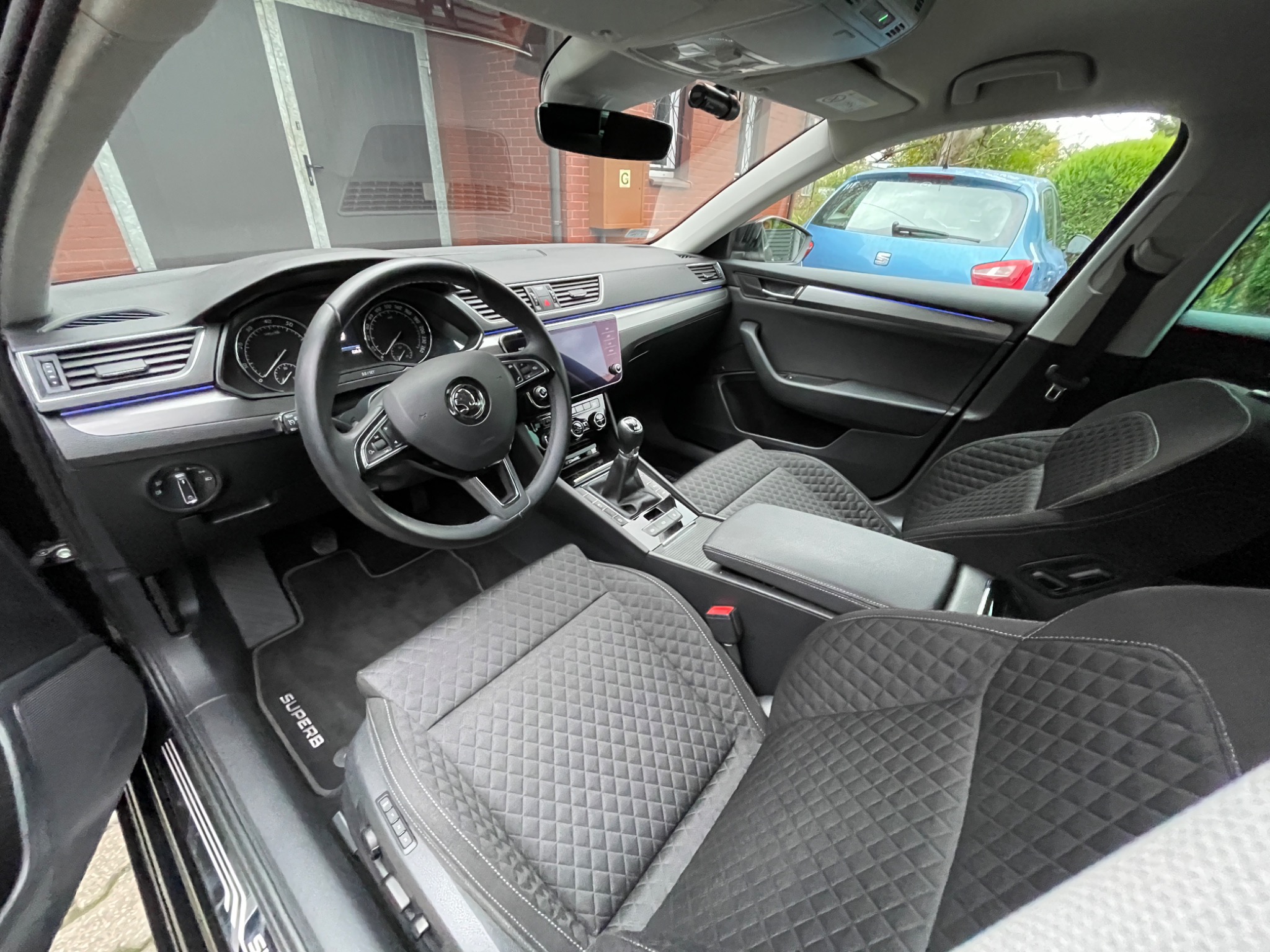 Wnętrze samochodu Skoda Superb z czarną tapicerką w romby, widok z przodu na deskę rozdzielczą, kierownicę i fotele, na podłodze dywanik z napisem 'Superb'.