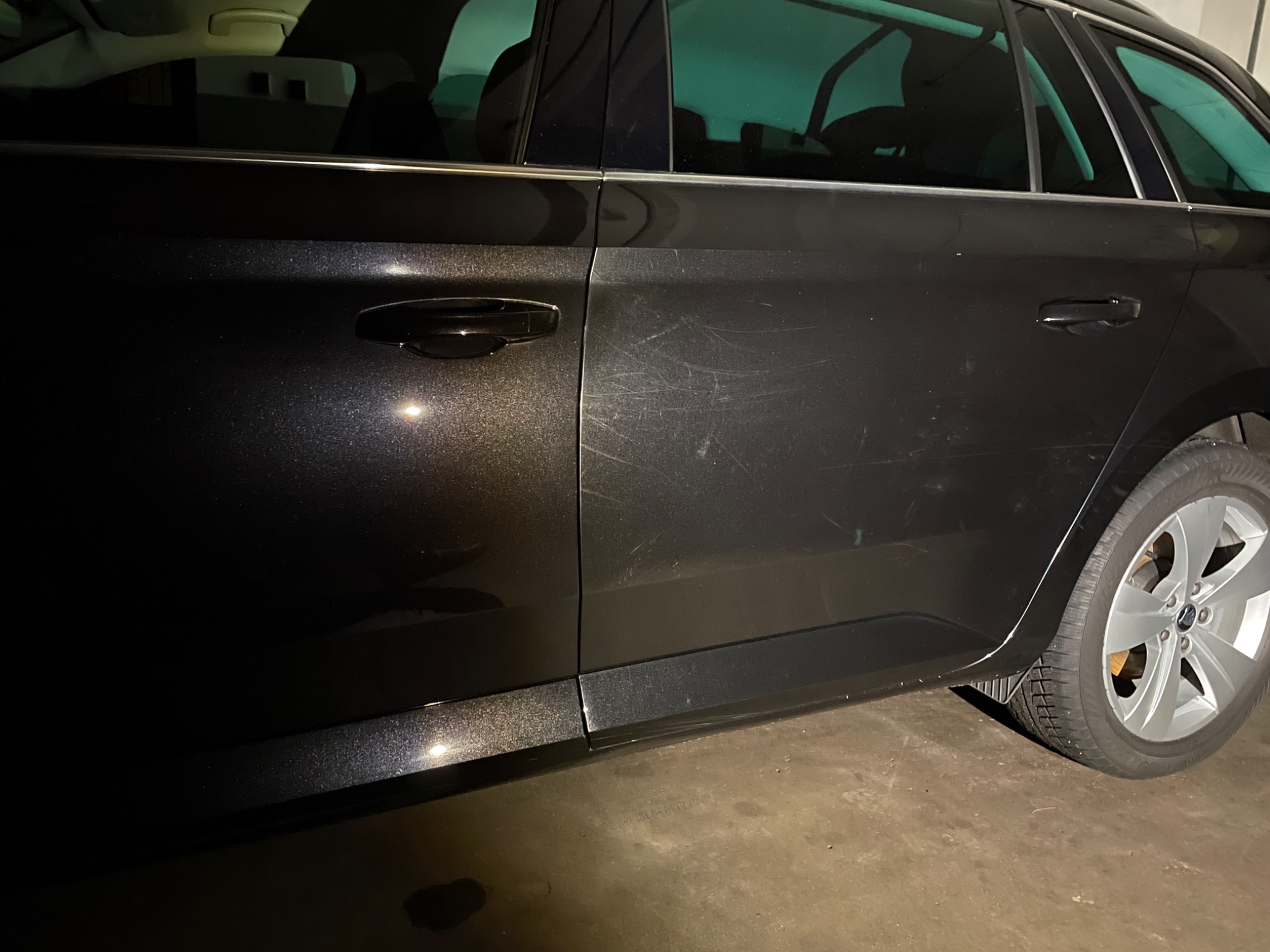 Ciemny samochód kombi z widocznymi zarysowaniami na bocznych drzwiach, opona z felgą aluminiową, sfotografowany w słabym oświetleniu.