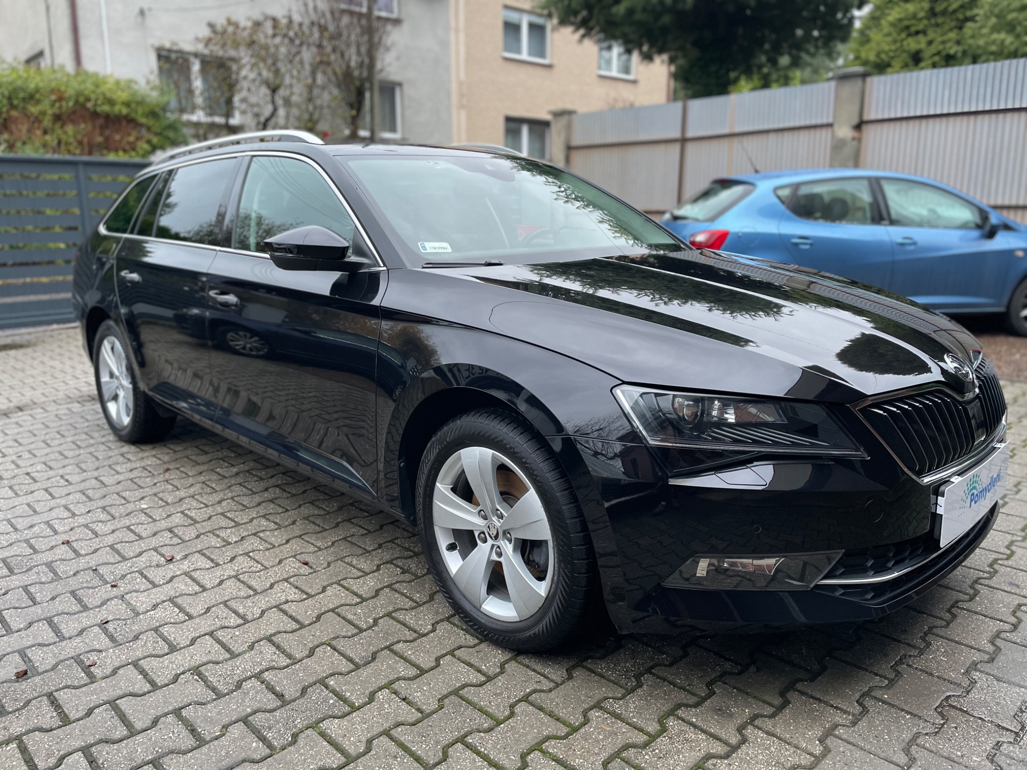 Czarny samochód kombi marki Skoda Superb zaparkowany na brukowanej nawierzchni, z widocznym niebieskim samochodem w tle i budynkiem mieszkalnym.