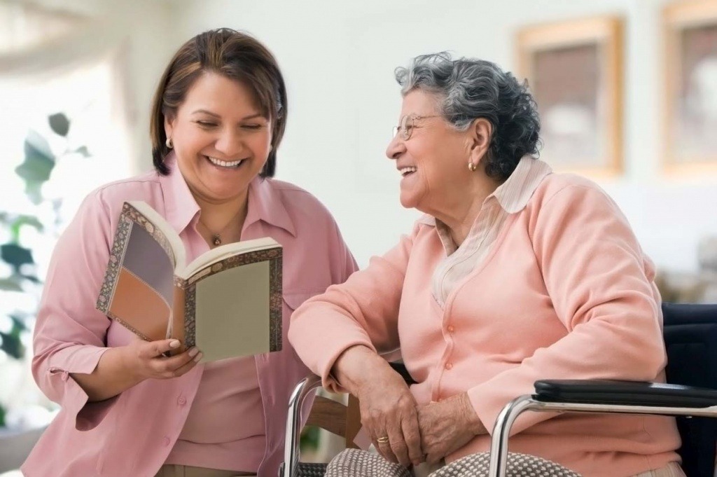 Uśmiechnięta kobieta czyta książkę starszej pani na wózku inwalidzkim w jasnym pokoju.