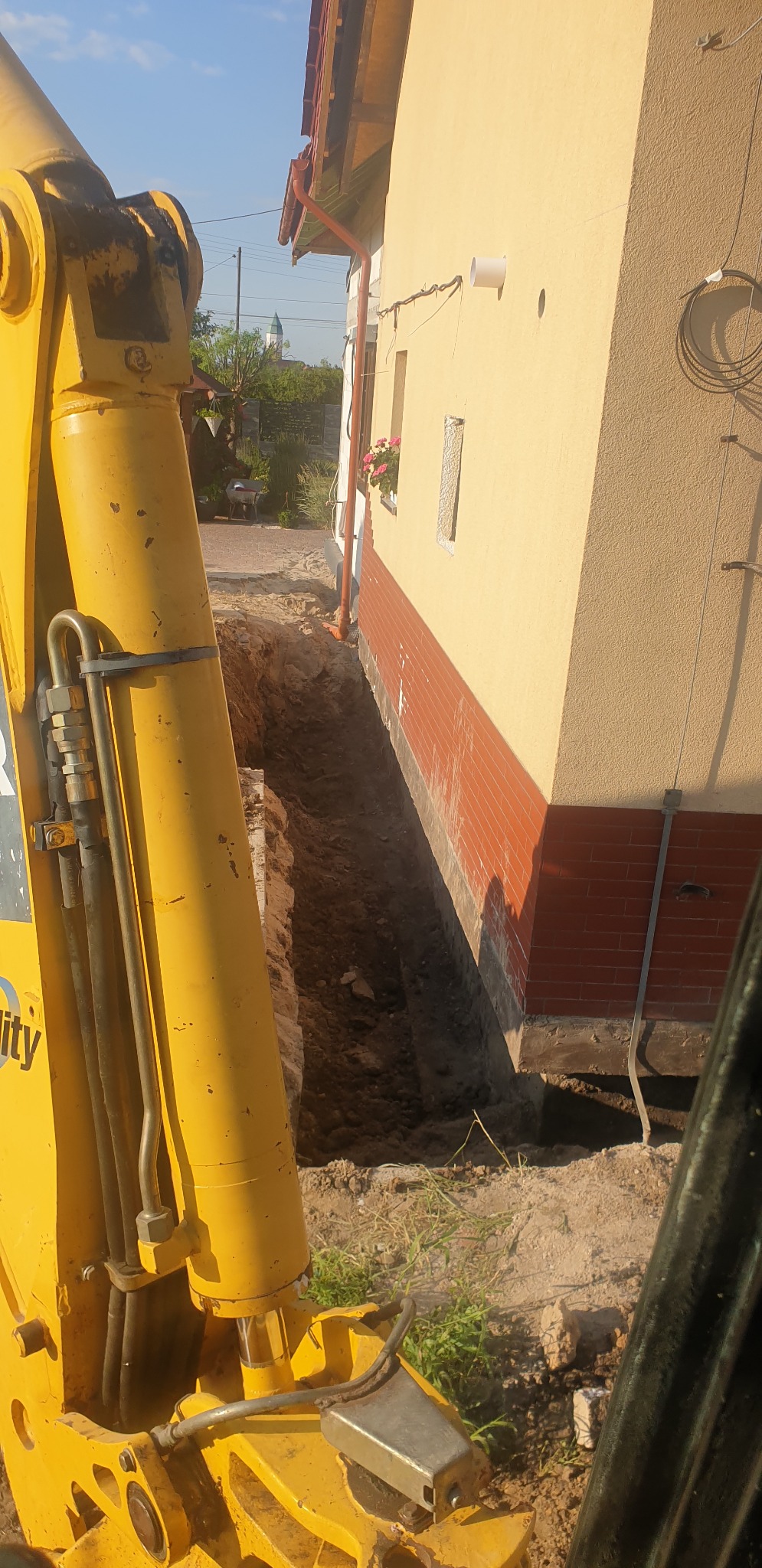 Żółta koparka przy wykopie wzdłuż ściany budynku z ceglaną podmurówką, widok z kabiny operatora, w tle zieleń i fragment wieży kościoła.